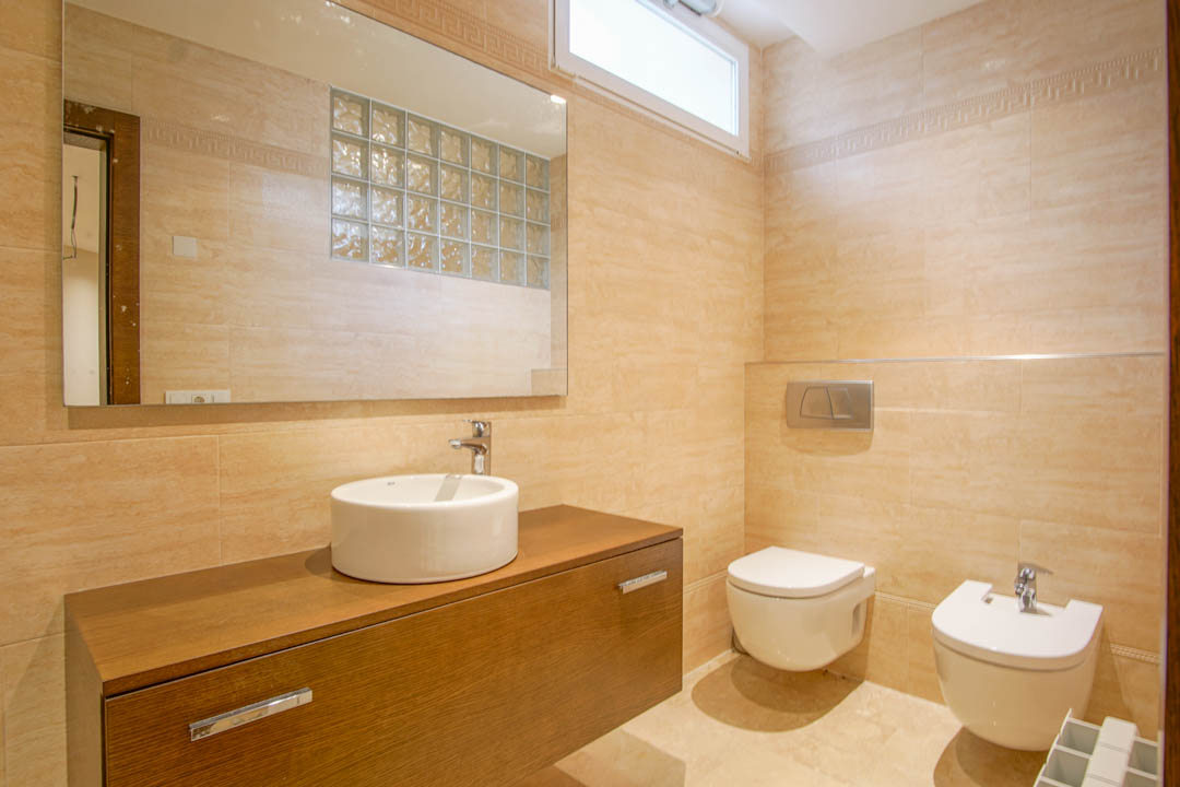 Salle d'eau carrelée en beige avec meuble suspendu en bois, vasque à poser, sanitaires muraux et miroir reflétant une paroi en briques de verre décoratives.