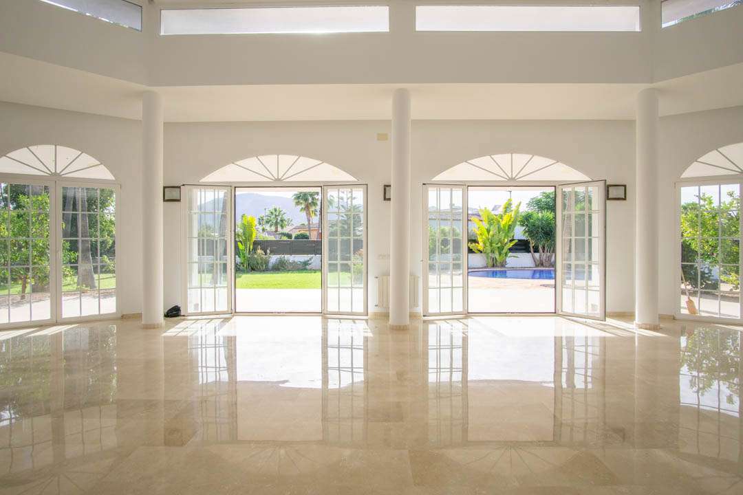 Vaste salon de style colonial avec sols en marbre poli, colonnes blanches et grandes portes-fenêtres cintrées offrant un accès direct au jardin et à la piscine.