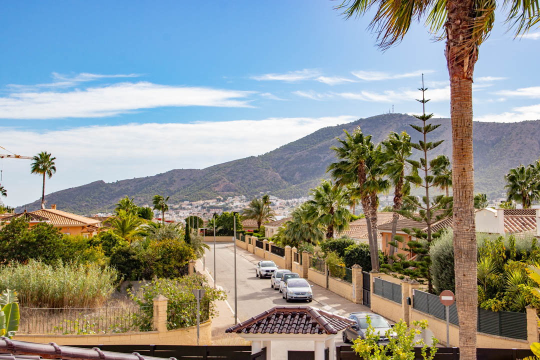 Vue dégagée depuis l'étage supérieur sur les environs résidentiels, montrant la rue bordée de palmiers et la silhouette de la montagne sous un ciel bleu.
