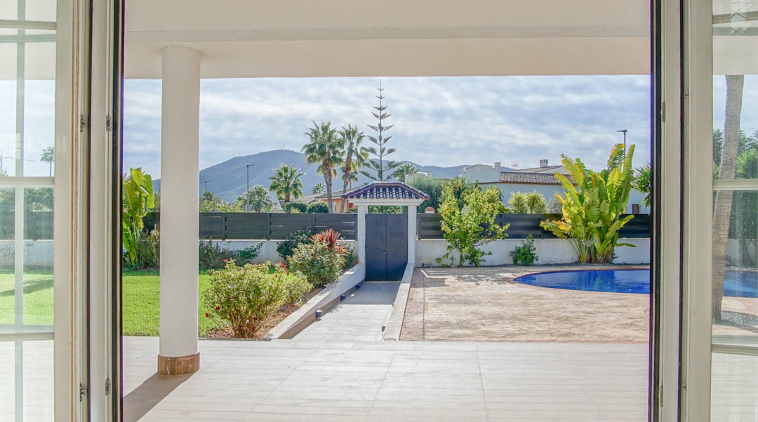 Vue depuis l'intérieur sur la terrasse couverte et le jardin, avec une piscine privée et des montagnes en arrière-plan à travers de larges portes vitrées.