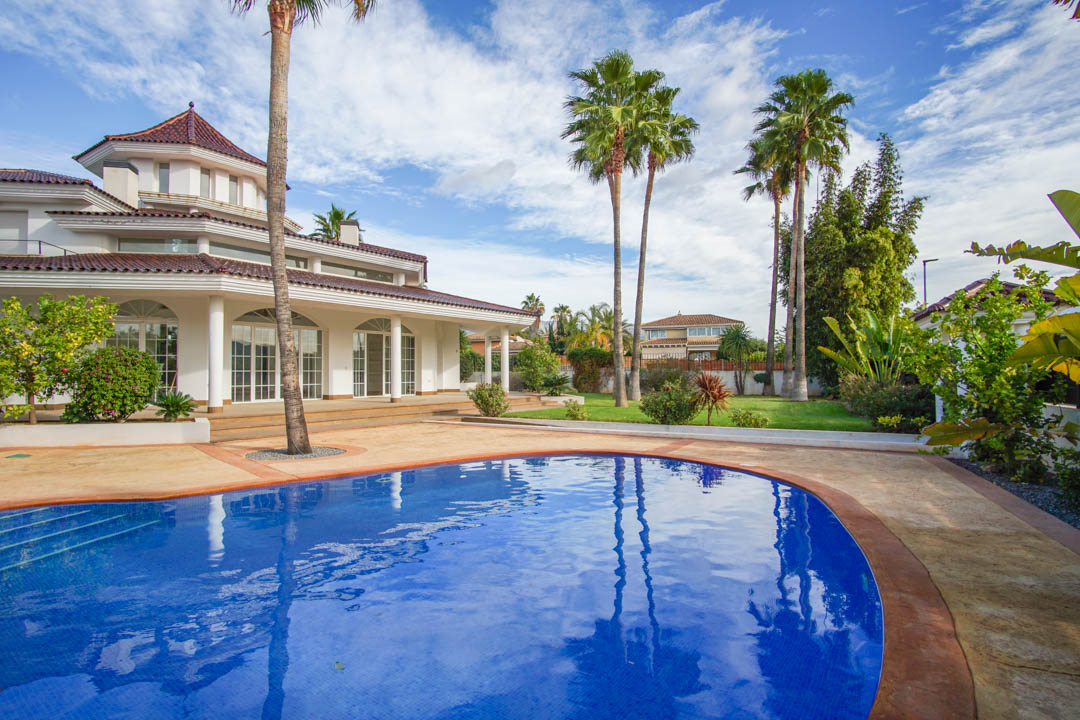 Vue extérieure de la villa coloniale avec piscine incurvée, jardin arboré de palmiers et façade blanche aux larges baies vitrées et colonnes.