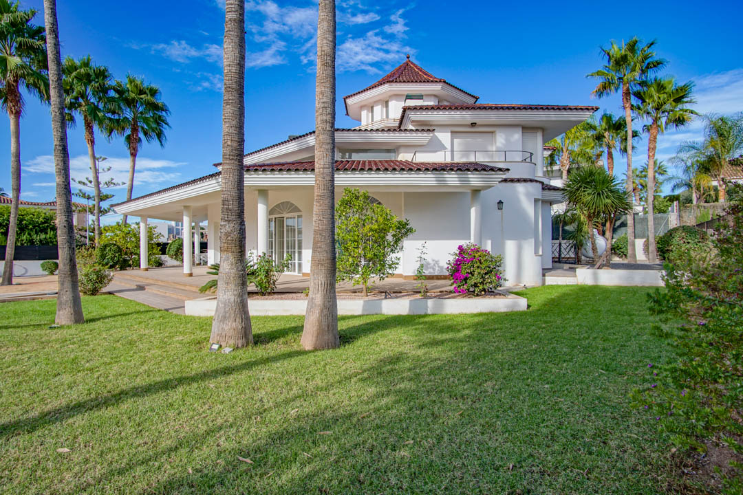 Vue extérieure de la villa coloniale blanche avec jardin paysager, palmiers matures et terrasses couvertes soutenues par des colonnes élégantes.
