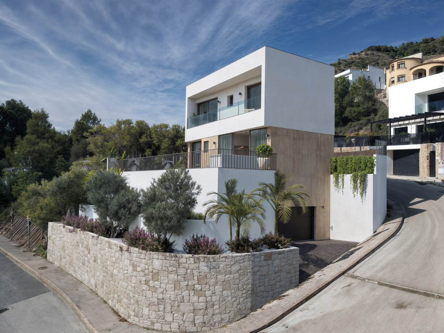 Villa moderne sur trois niveaux avec façade en pierre, terrasses aux garde-corps en verre et garage en sous-sol, nichée dans un paysage vallonné.