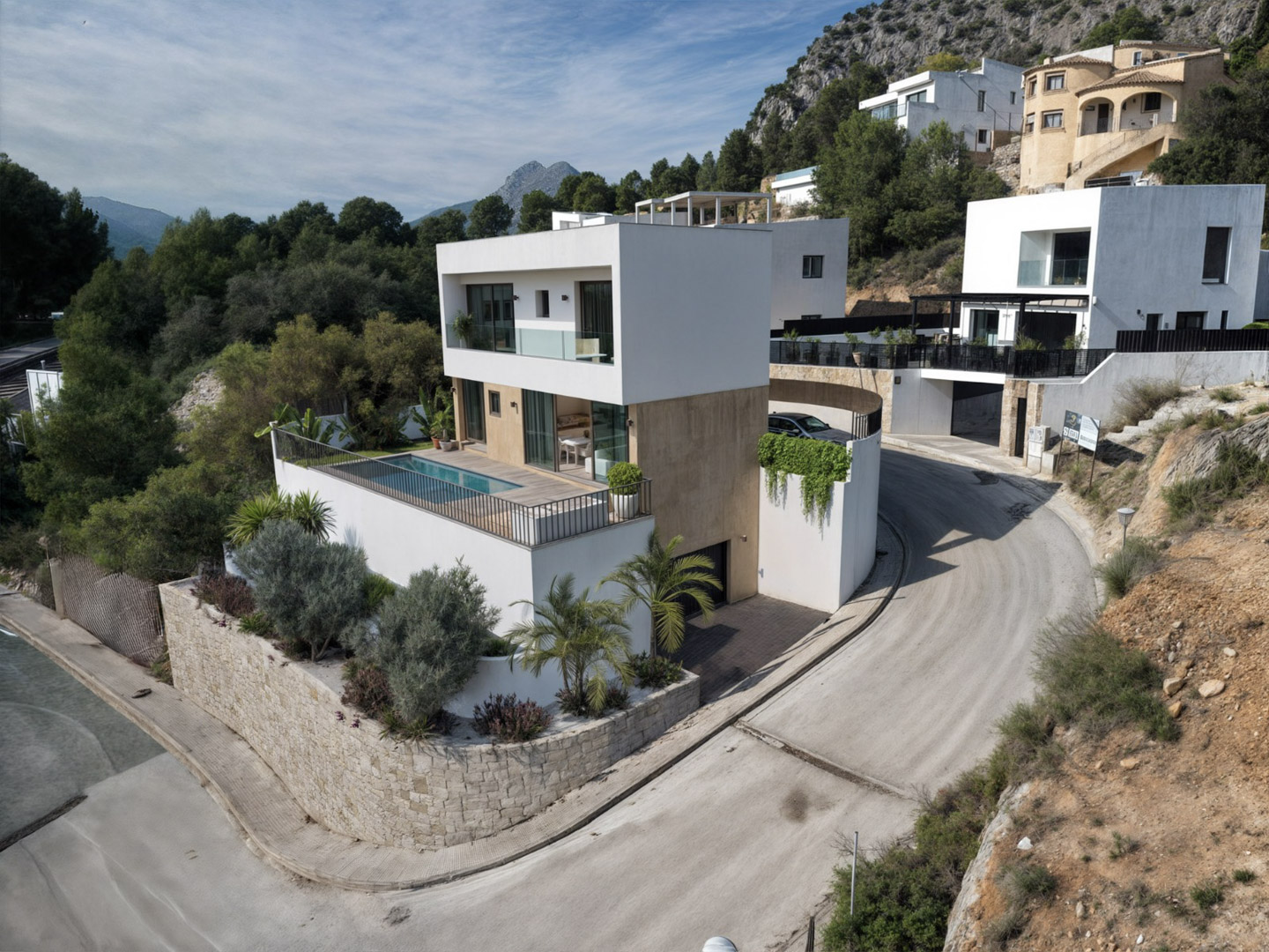Vue extérieure de la villa moderne alliant blanc et pierre, avec terrasse surélevée, piscine privée et vue sur les montagnes environnantes d'Alcalalí.