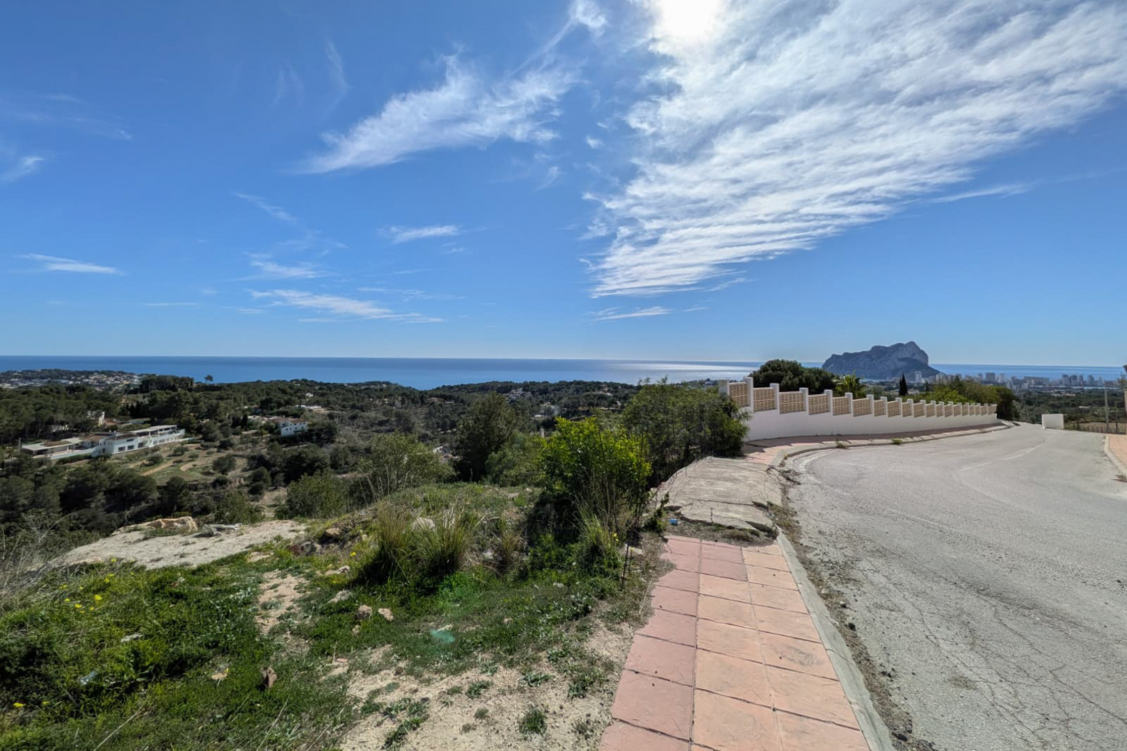 Vue panoramique depuis la route d'accès, mettant en valeur la côte de Benissa, la mer Méditerranée et la silhouette du Peñón de Ifach à l'horizon.