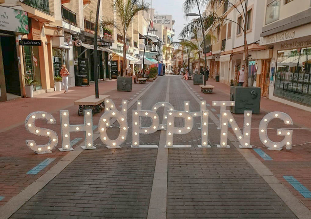 Rue commerçante piétonne au centre de Moraira, bordée de palmiers et de boutiques, avec une grande enseigne lumineuse décorative au premier plan.
