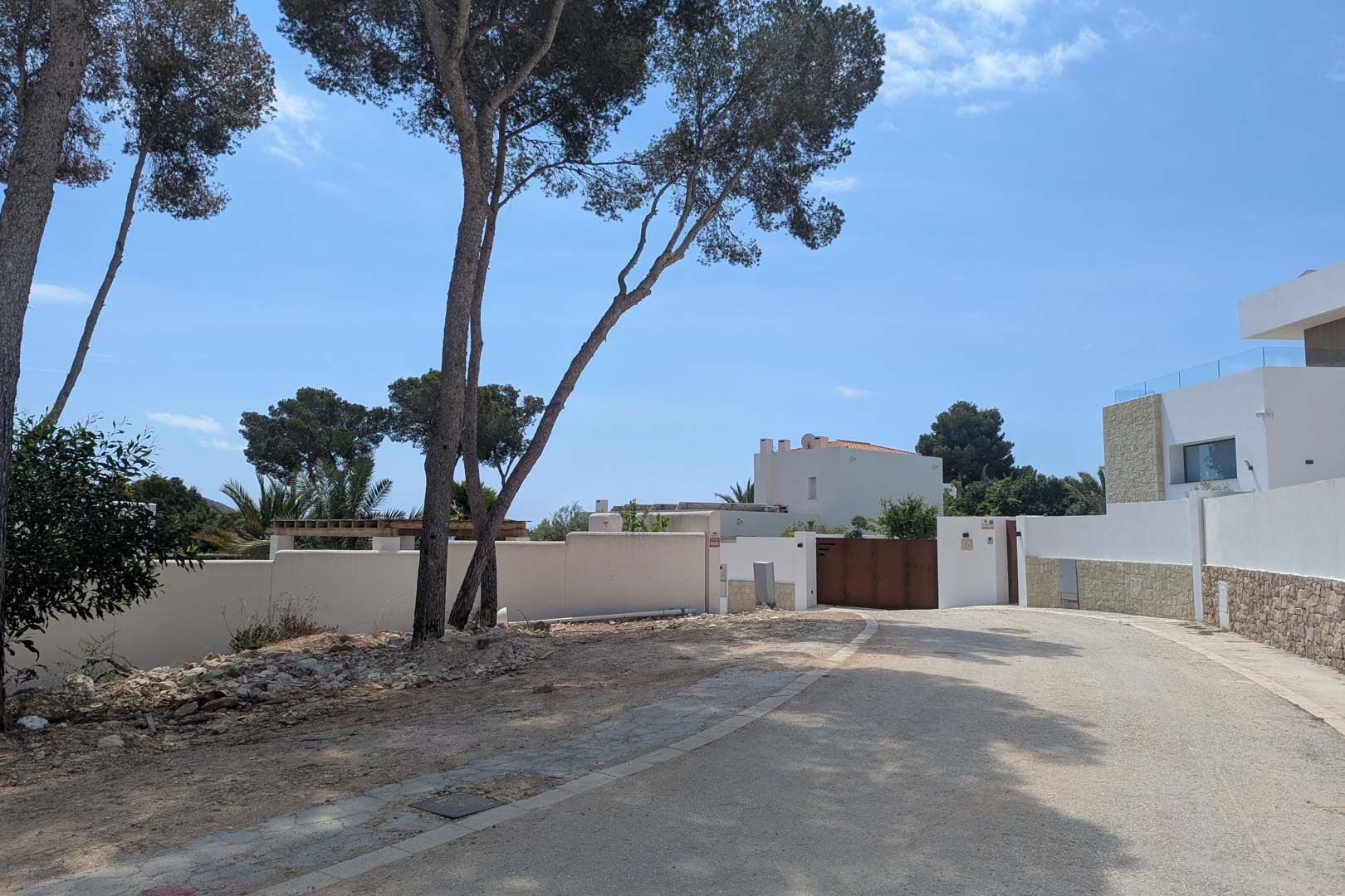 Vue de la rue d'accès dans un quartier résidentiel calme de Moraira, avec des pins matures et des murs d'enceinte blancs de style méditerranéen.
