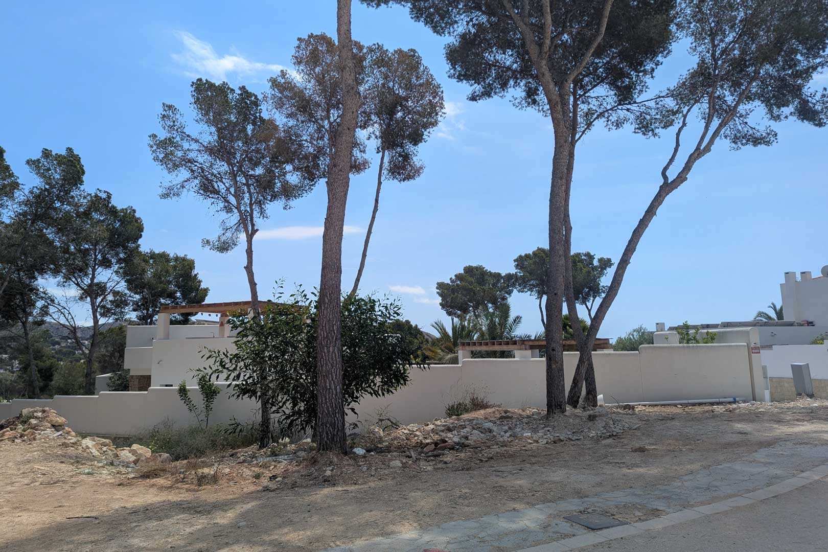 Vue du terrain à bâtir avec des pins matures et des murs d'enceinte blancs, prêt pour la construction de la villa de style ibizien à Moraira.