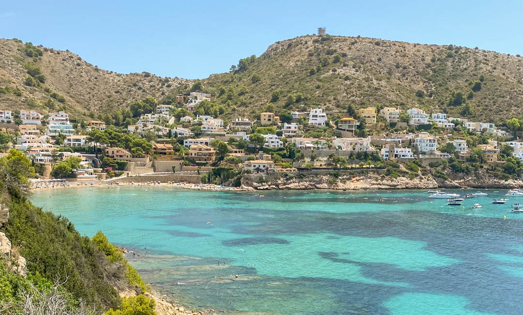 Vue panoramique de la plage d'El Portet à Moraira, avec ses eaux turquoise, ses bateaux au mouillage et ses villas blanches nichées sur la colline côtière.