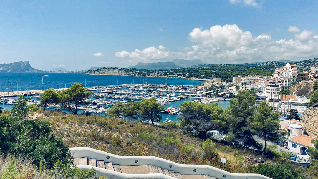 Vue panoramique sur le port de plaisance de Moraira et le Peñón de Ifach depuis un escalier blanc de style méditerranéen bordé de végétation.