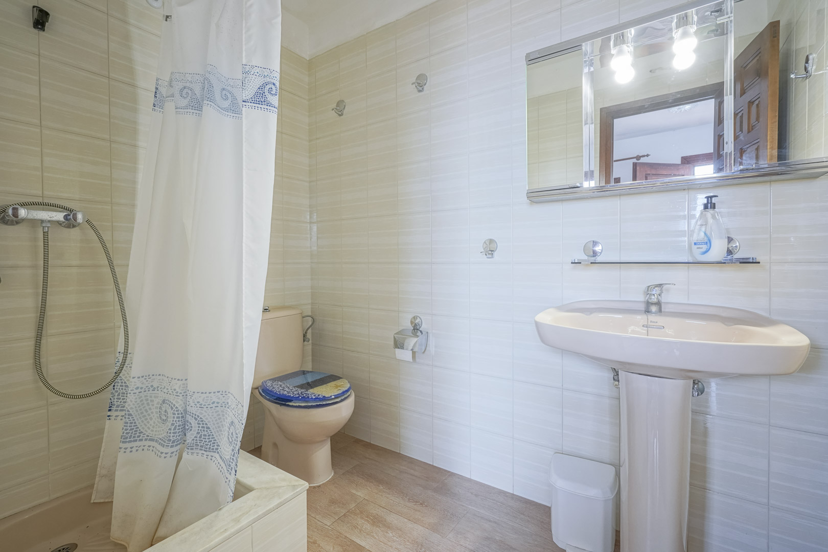 Salle de bain avec carrelage clair, lavabo sur pied rose et rideau de douche à motifs bleus.
