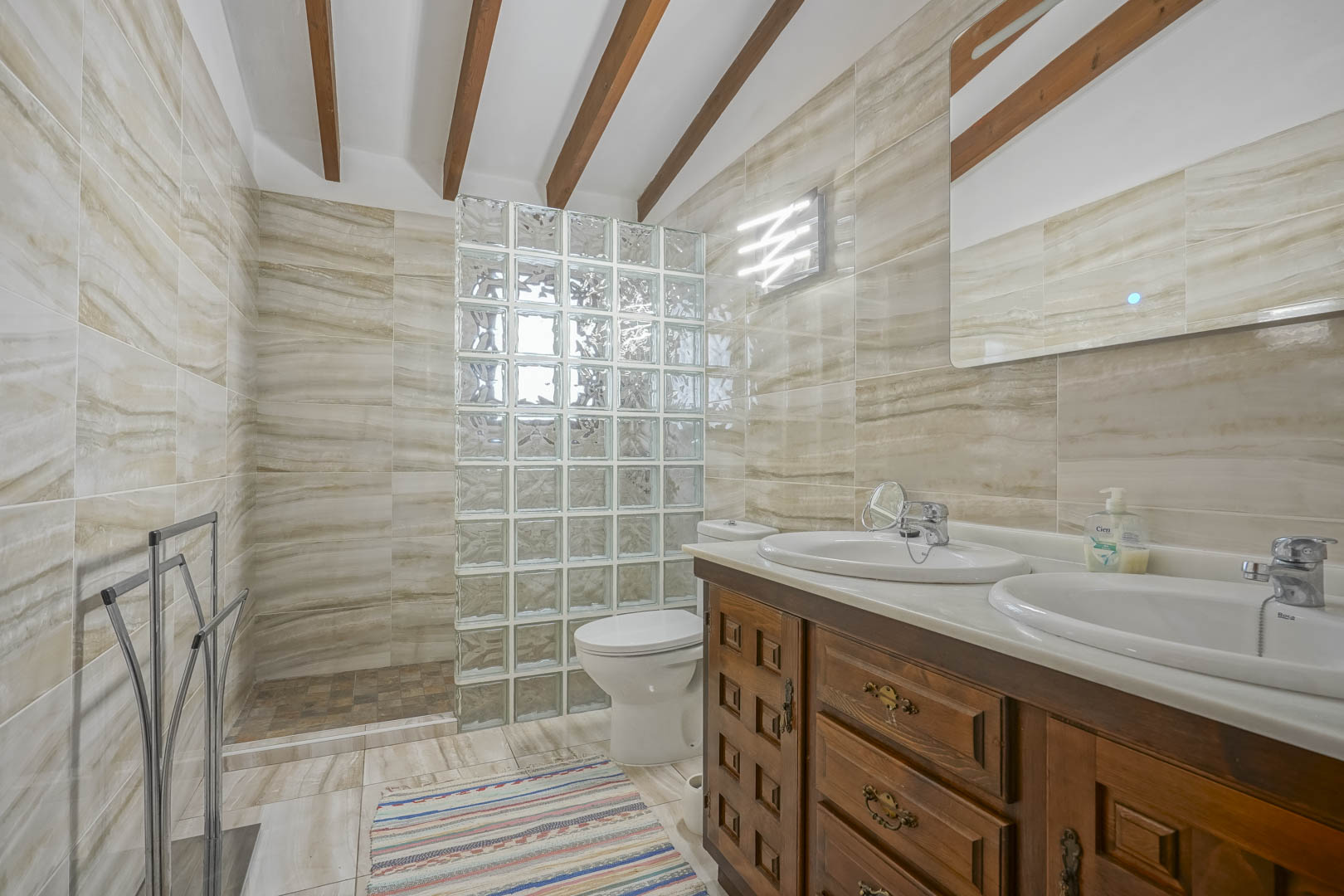 Salle de bain avec paroi de douche en pavés de verre, meuble-lavabo en bois avec double vasque et carrelage veiné.