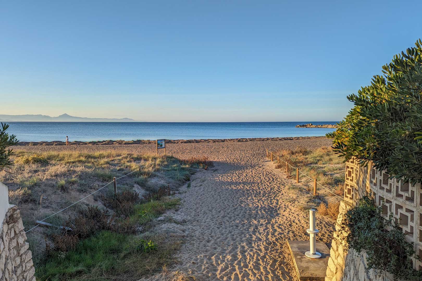 Vue sur une plage de sable avec un chemin délimité, de la végétation côtière et la mer bleue sous un ciel clair.
