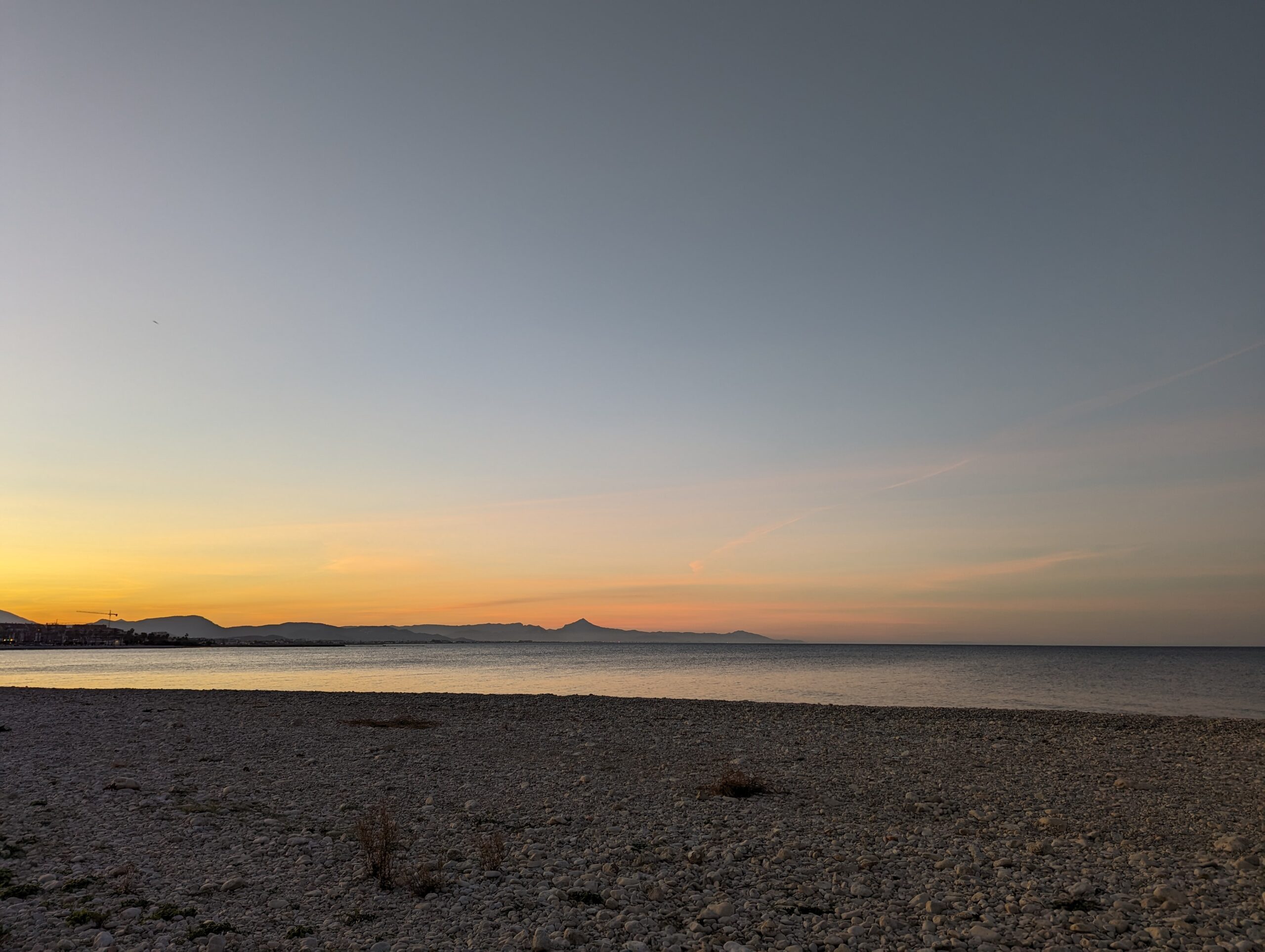 Vue sur la plage de galets au coucher du soleil avec des montagnes au loin.
