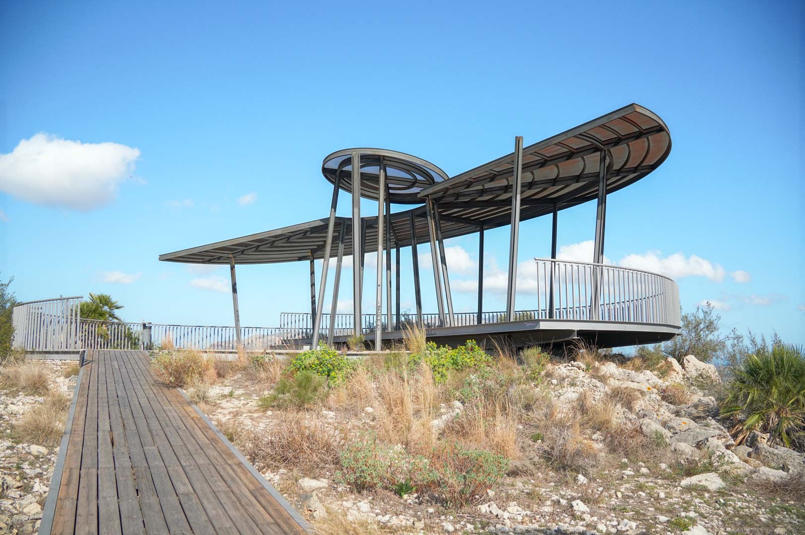Belvédère au design moderne avec structure métallique et toit incurvé, accessible par une passerelle en bois au cœur d'un paysage méditerranéen préservé.
