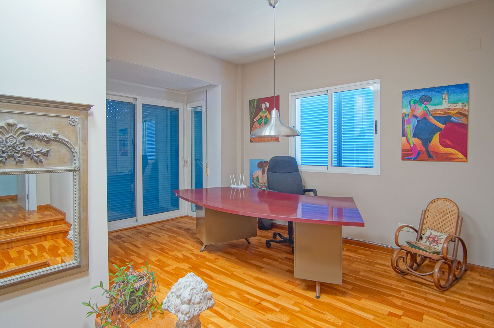 Bureau lumineux avec parquet, un grand bureau incurvé bordeaux et des fenêtres équipées de volets bleus traditionnels apportant une lumière naturelle.