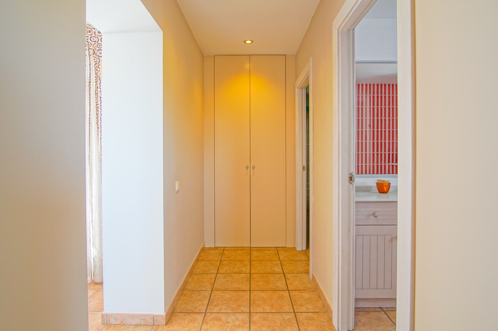 Couloir avec sol en grès cérame, comprenant un placard intégré toute hauteur et offrant un accès à une salle de bain aux carreaux muraux rayés rouges et blancs.