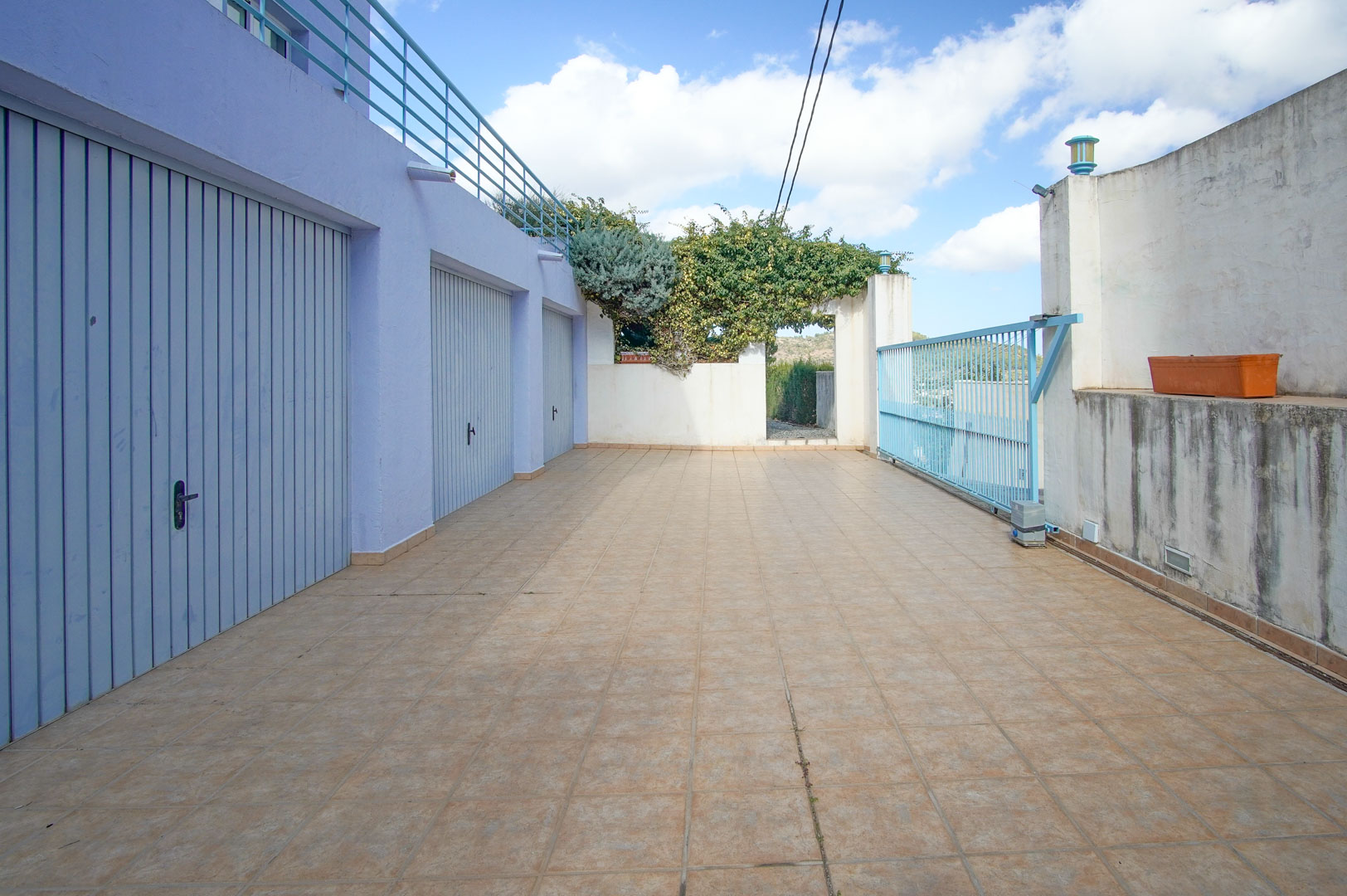 Entrée inférieure avec trois garages privés aux portes bleu clair, une large allée carrelée et un portail coulissant métallique sous la terrasse supérieure.