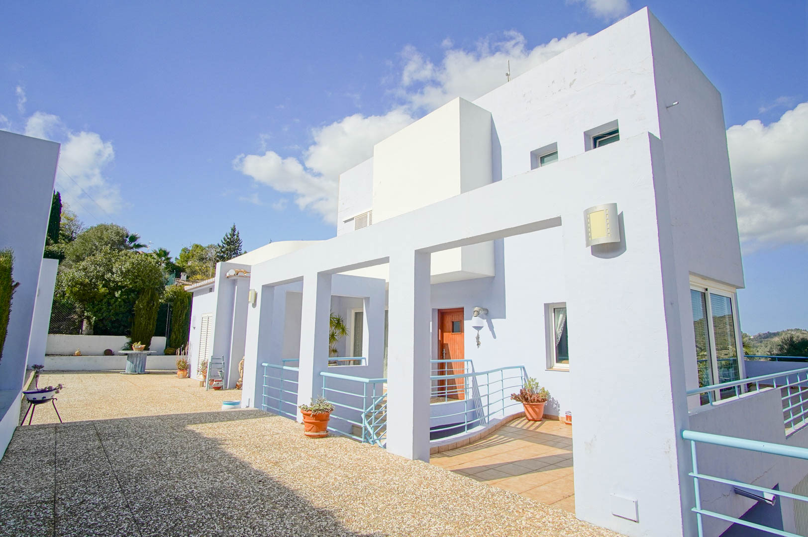 Façade moderne aux tons clairs avec accents bleu ciel, vaste terrasse en gravier et entrée principale avec porte en bois et garde-corps métalliques.
