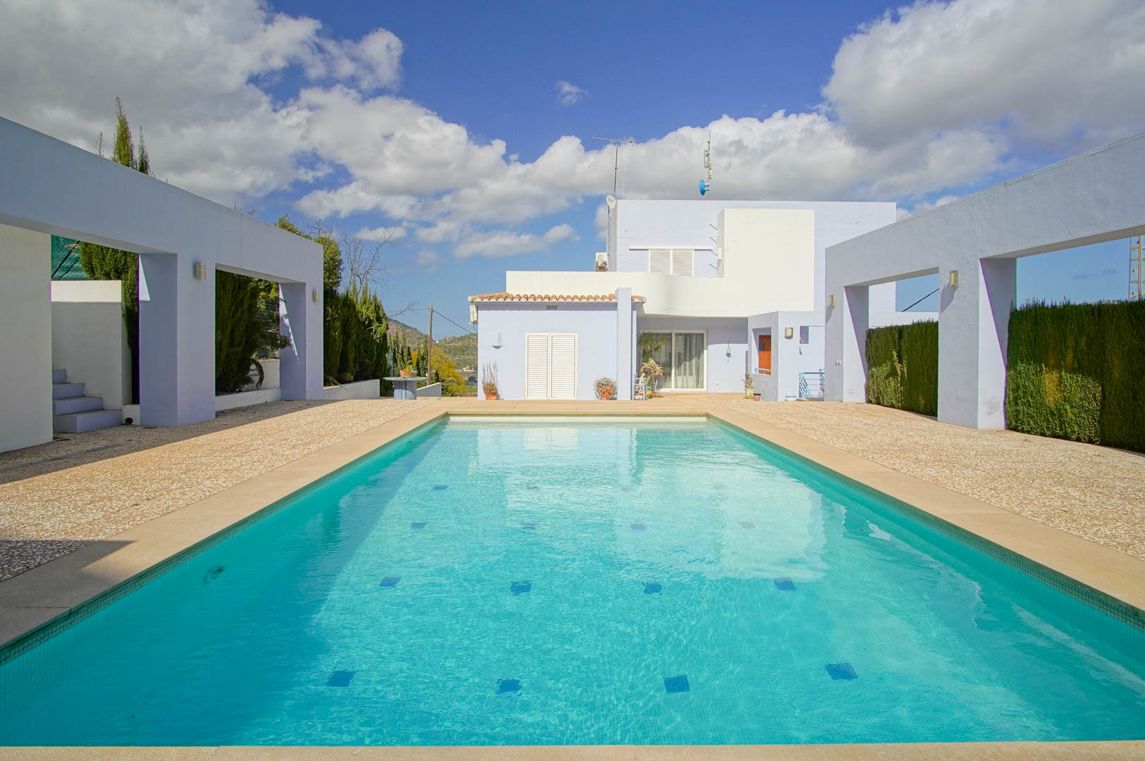 Piscine rectangulaire avec mosaïque turquoise, entourée d'une vaste terrasse en gravier et de portiques modernes devant la façade de la villa.