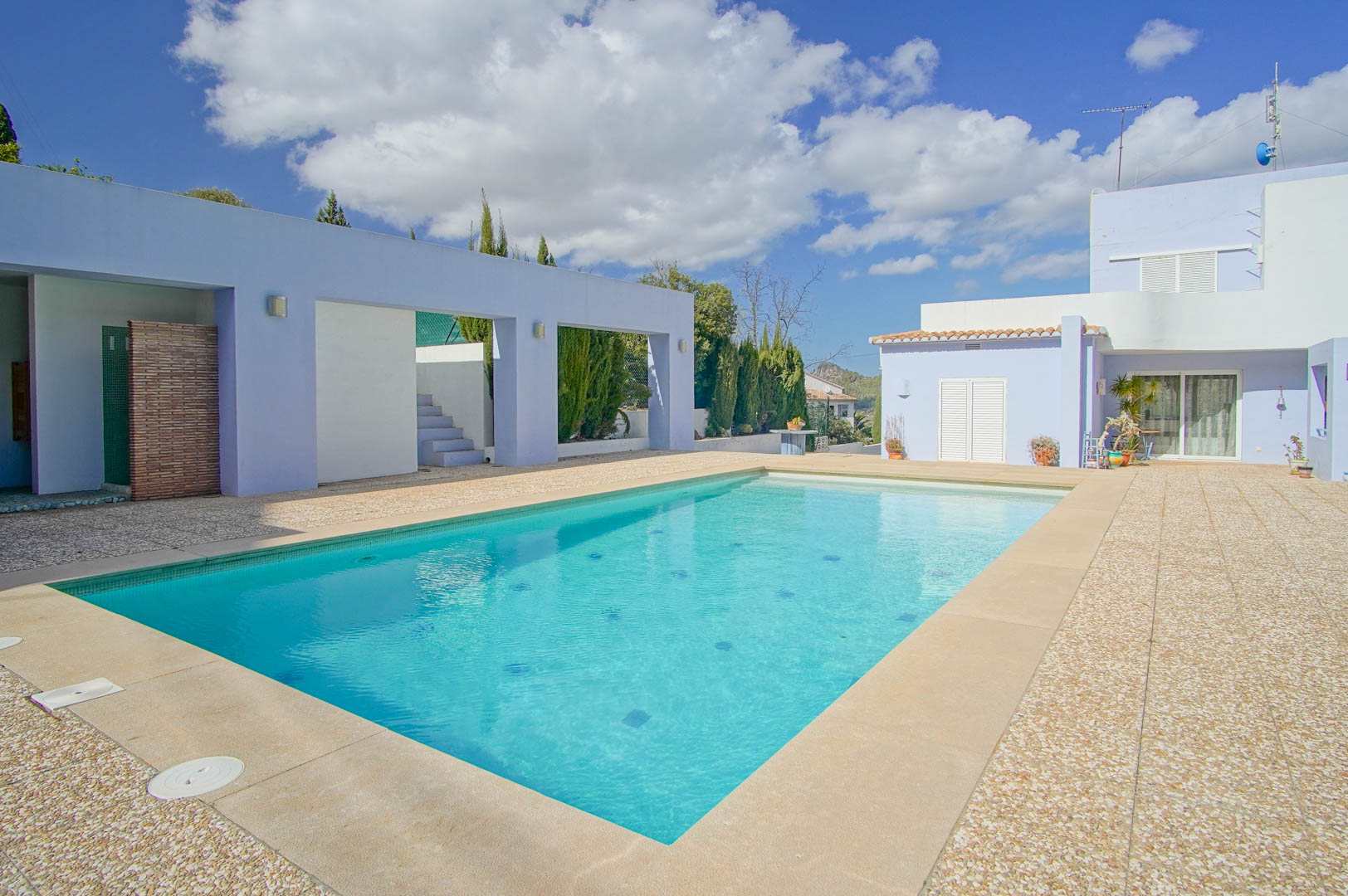 Piscine rectangulaire entourée d'une terrasse en gravier et pierre, bordée de murs aux tons lavande et d'une architecture moderne aux lignes épurées.