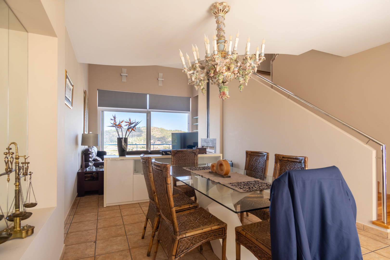 Salle à manger avec table en verre, chaises en osier et lustre ornemental. Une grande fenêtre offre une vue sur la montagne à côté de l'escalier à rampe métallique.
