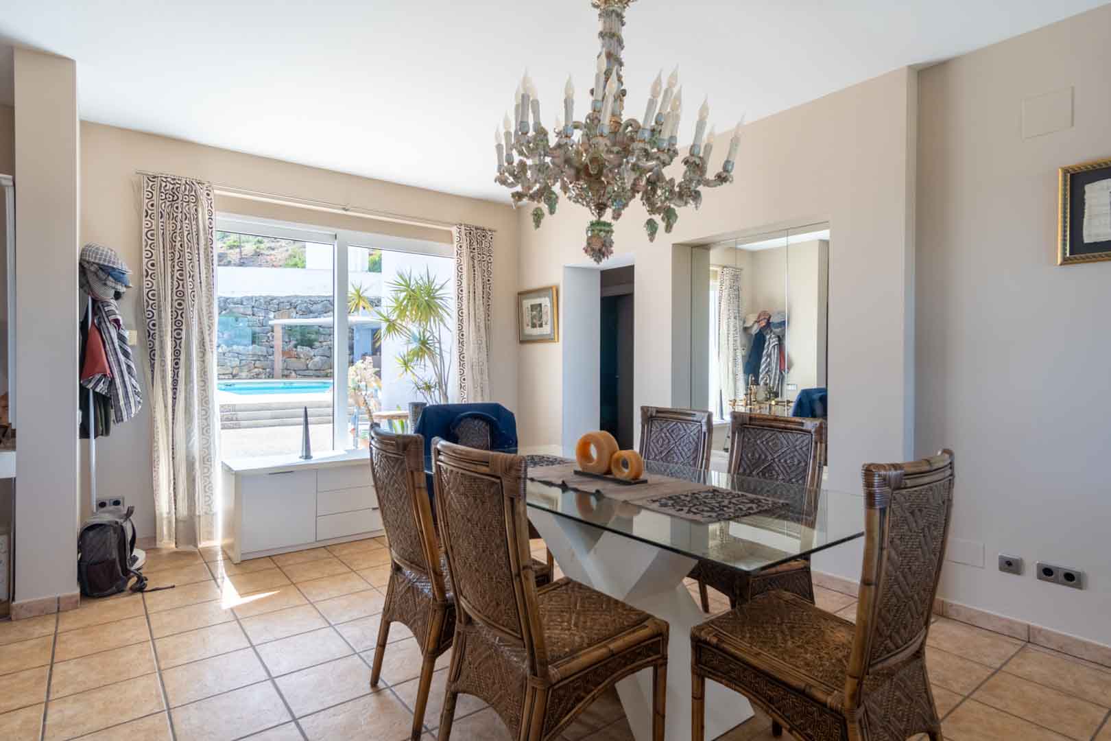 Salle à manger lumineuse avec table en verre, chaises en osier et lustre classique, offrant une vue dégagée sur la piscine extérieure grâce à de larges baies vitrées.