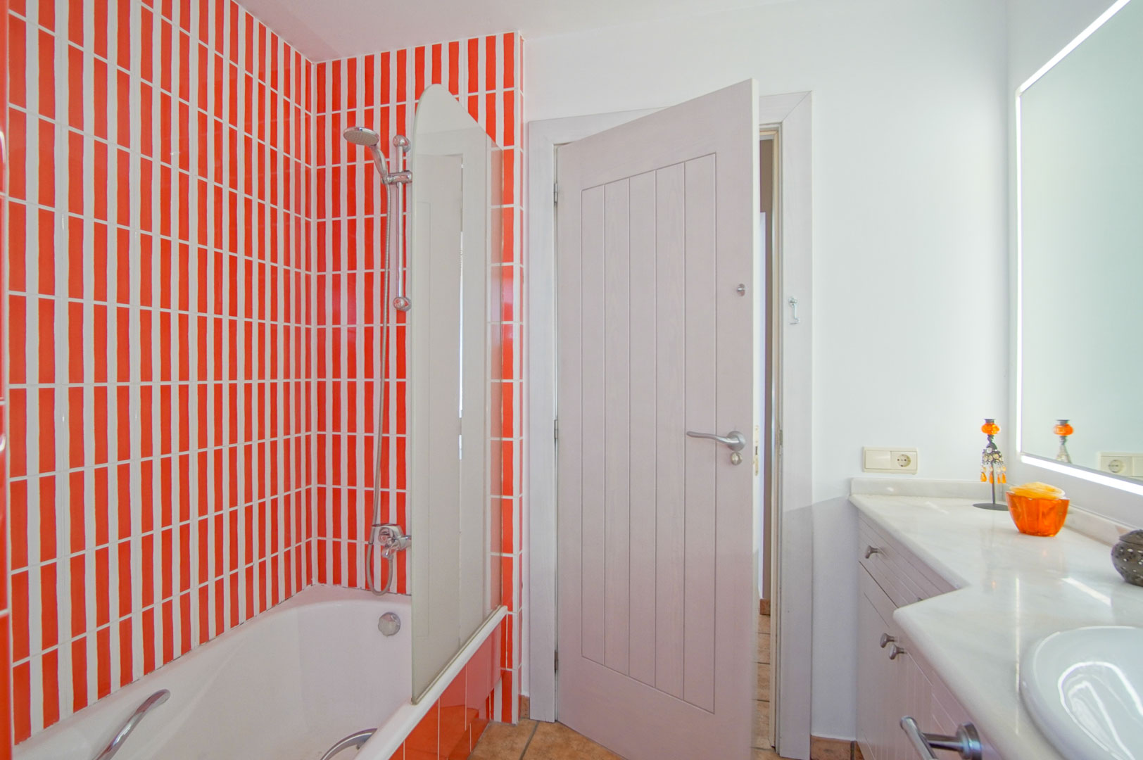 Salle de bain avec baignoire et paroi en verre, caractérisée par un carrelage vertical orange et blanc, un plan de travail en marbre et un miroir à éclairage intégré.