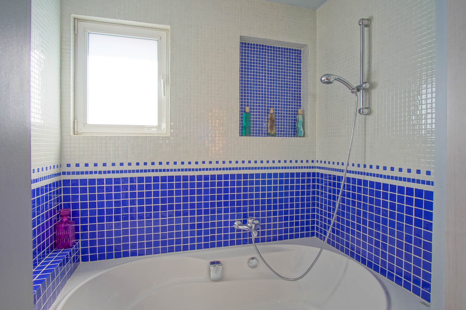 Salle de bain avec baignoire ovale et revêtement en mosaïque bleue et blanche. Elle dispose d'une niche intégrée pratique et d'une fenêtre offrant une luminosité naturelle.