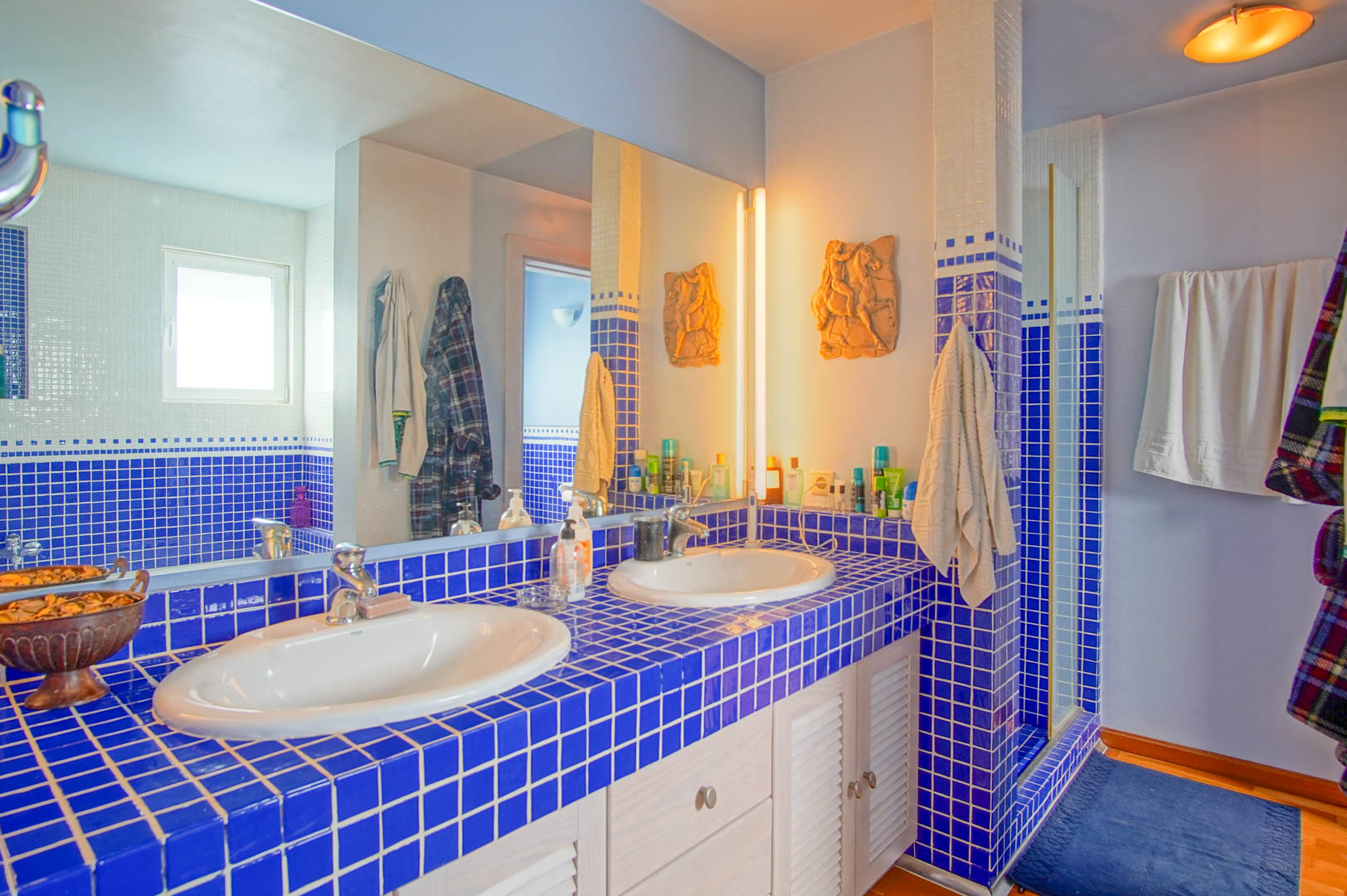 Salle de bain avec double vasque sur plan de travail en carreaux bleus. Comprend des meubles blancs à persiennes et un grand miroir reflétant la douche.
