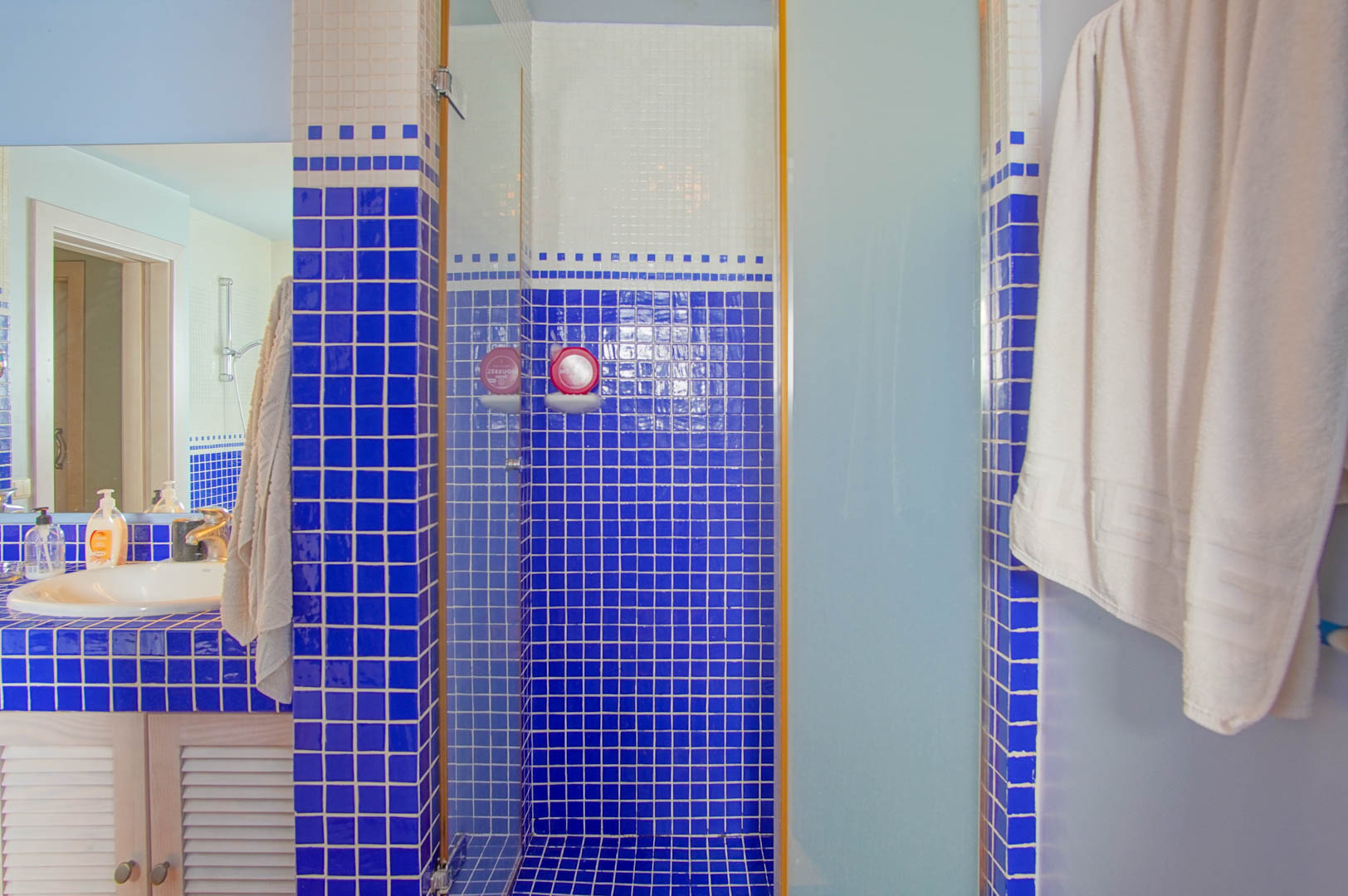 Salle de bain avec mosaïque bleue et blanche, douche à l'italienne avec paroi en verre et meuble vasque en bois aux portes persiennes.