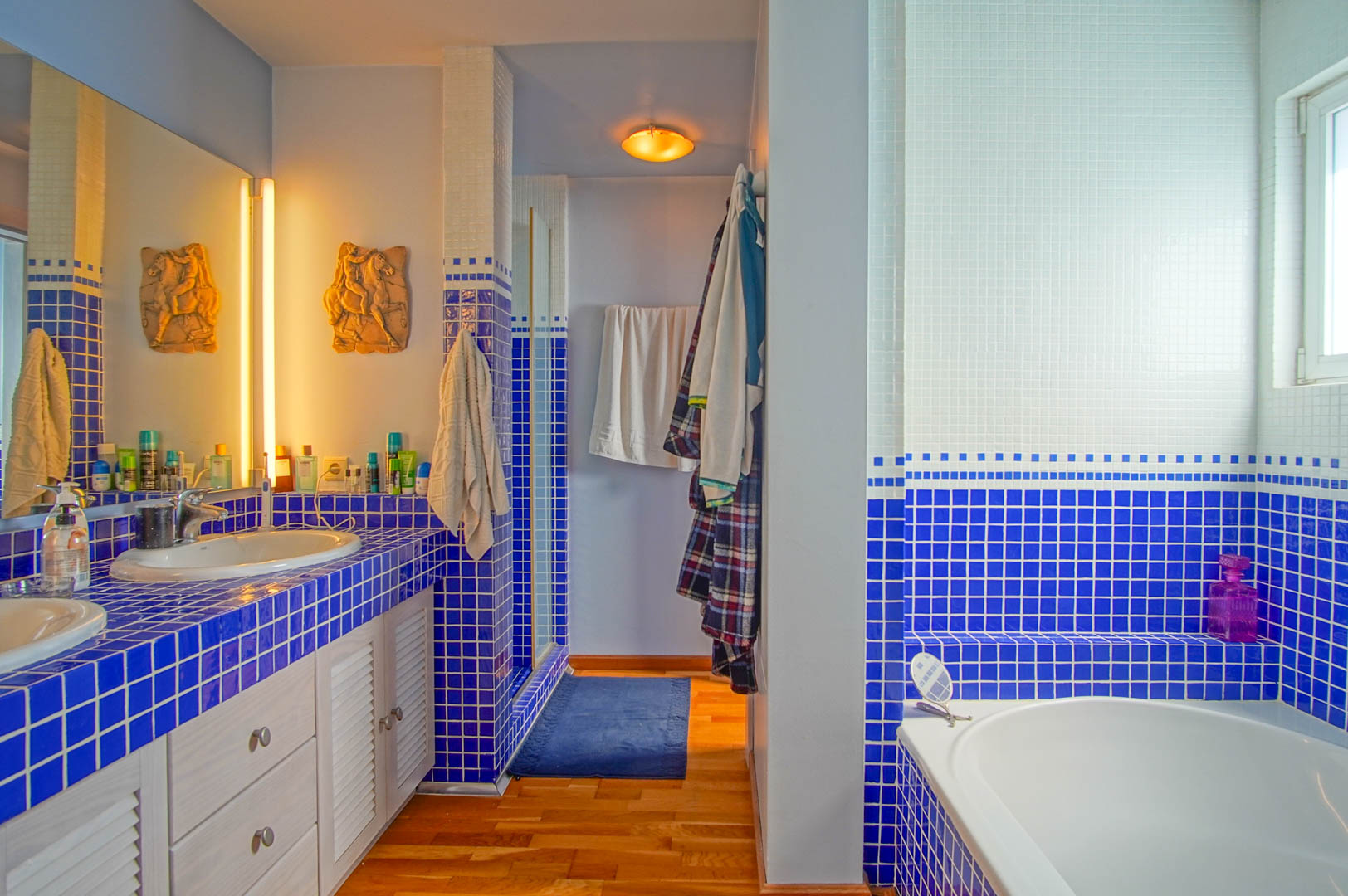 Salle de bain avec plan vasque en mosaïque bleue et blanche, double évier, parquet au sol et baignoire intégrée. L'espace dispose de rangements à persiennes blanches.