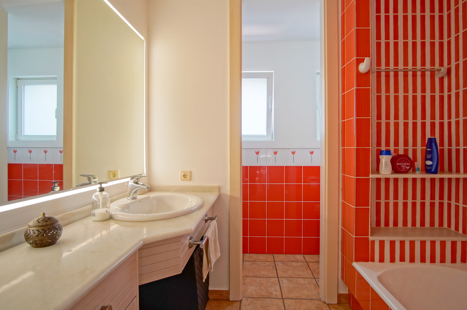 Salle de bain fonctionnelle avec plan de travail en marbre, miroir rétroéclairé et carrelage orange vif. Comprend une baignoire avec étagères intégrées et lumière naturelle.