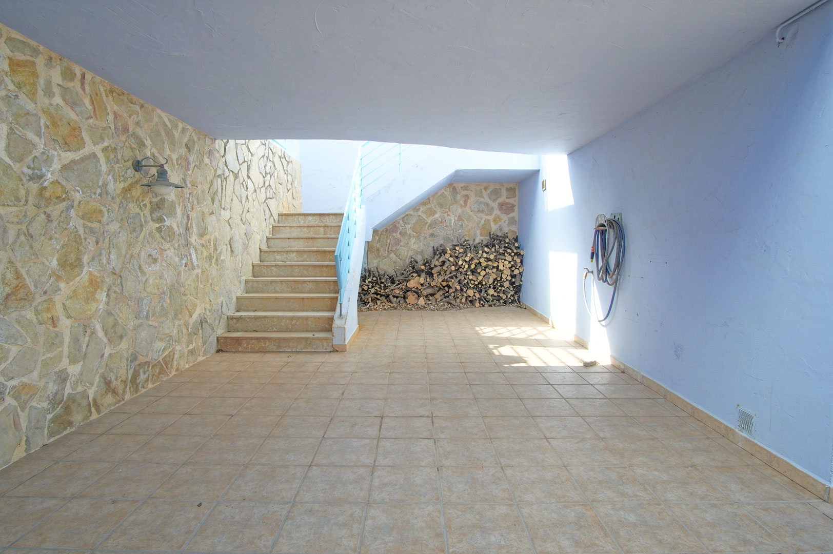 Terrasse couverte avec mur en pierre naturelle, sol en grès et espace de rangement pour le bois sous l'escalier extérieur menant à l'étage.