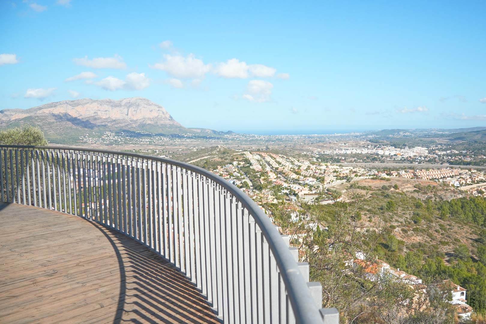 Terrasse incurvée avec parquet en bois et garde-corps métallique, offrant une vue panoramique sur la vallée, les montagnes et la mer Méditerranée.