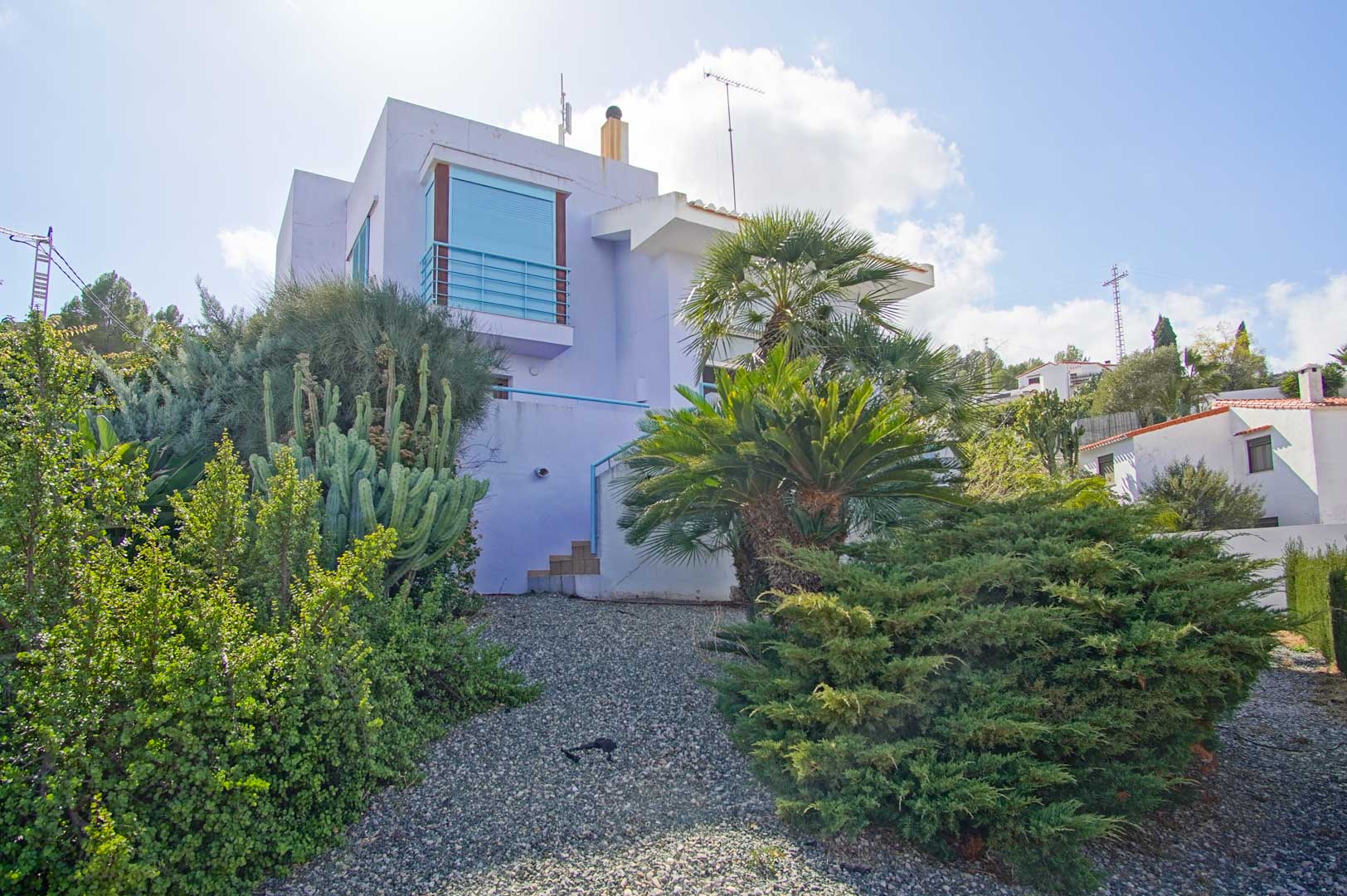 Vue extérieure de la façade aux tons lavande, intégrée dans un jardin méditerranéen avec cactus, palmiers et arbustes sur un terrain de gravier.