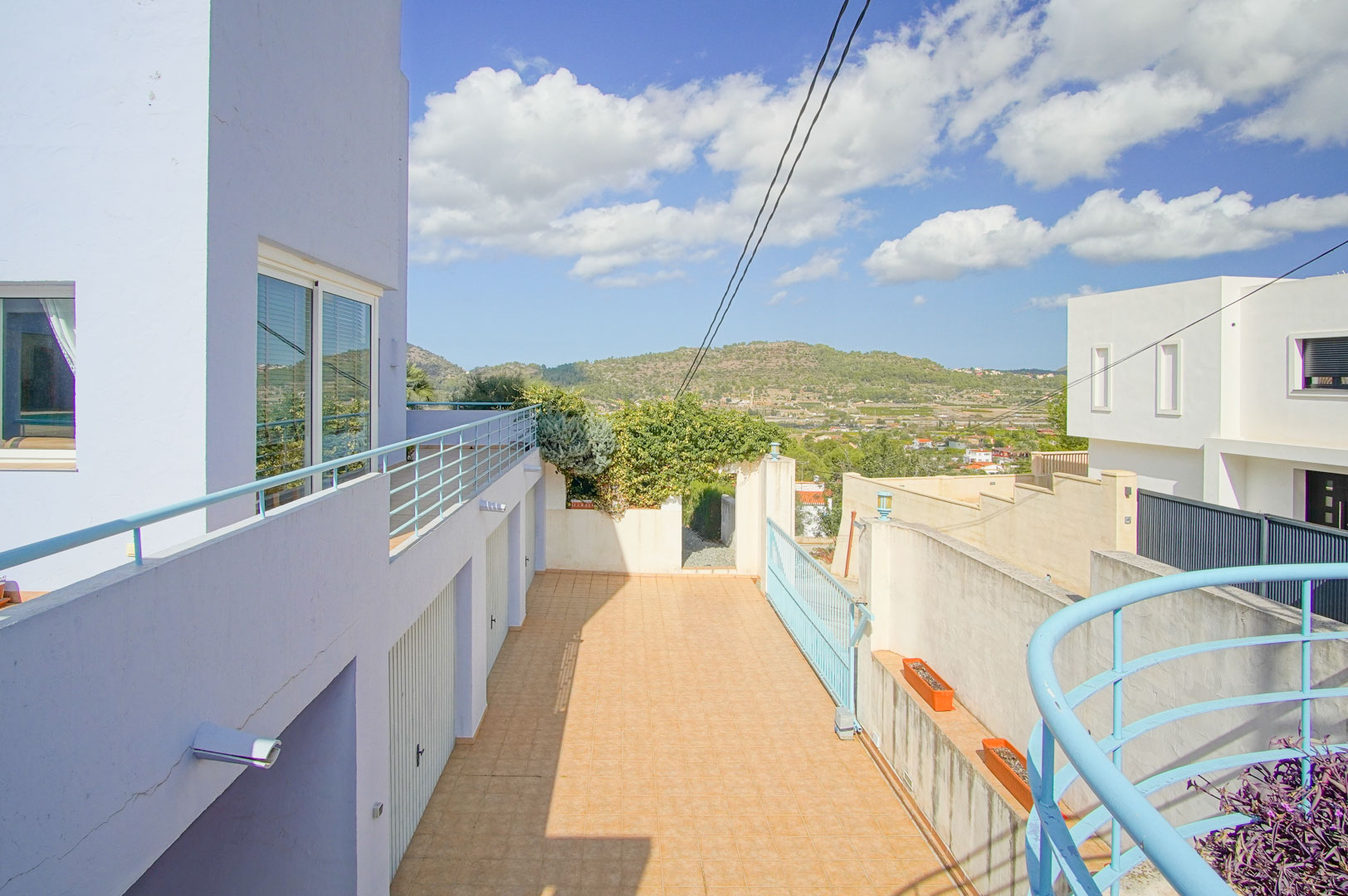 Vue extérieure de l'allée menant aux trois garages privés, avec carrelage au sol et garde-corps bleus, offrant une vue dégagée sur les montagnes.