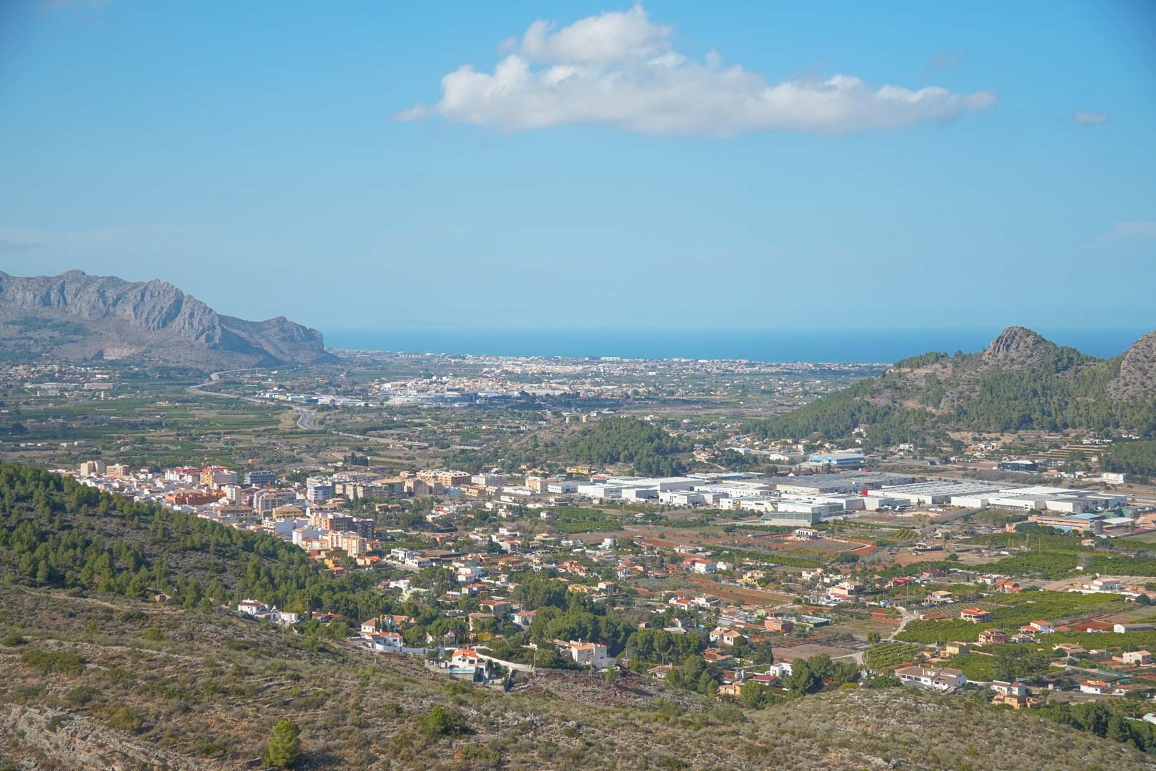 Vue panoramique surélevée montrant la vallée de Pedreguer, les chaînes de montagnes et la mer Méditerranée à l'horizon sous un ciel bleu clair.