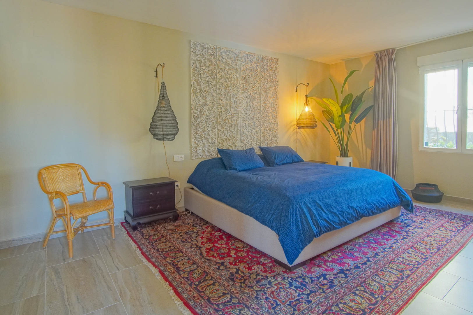 Chambre avec panneau mural sculpté, suspensions en osier et tapis oriental. La pièce dispose de sols en grès cérame et d'une luminosité naturelle constante.