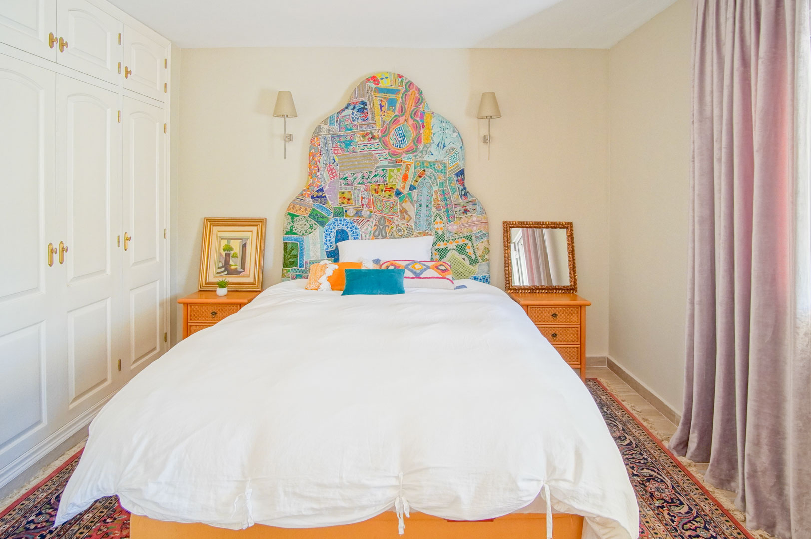 Chambre avec une tête de lit artisanale colorée, des armoires encastrées blanches et des tables de chevet en bois. Un tapis oriental complète cet espace lumineux.