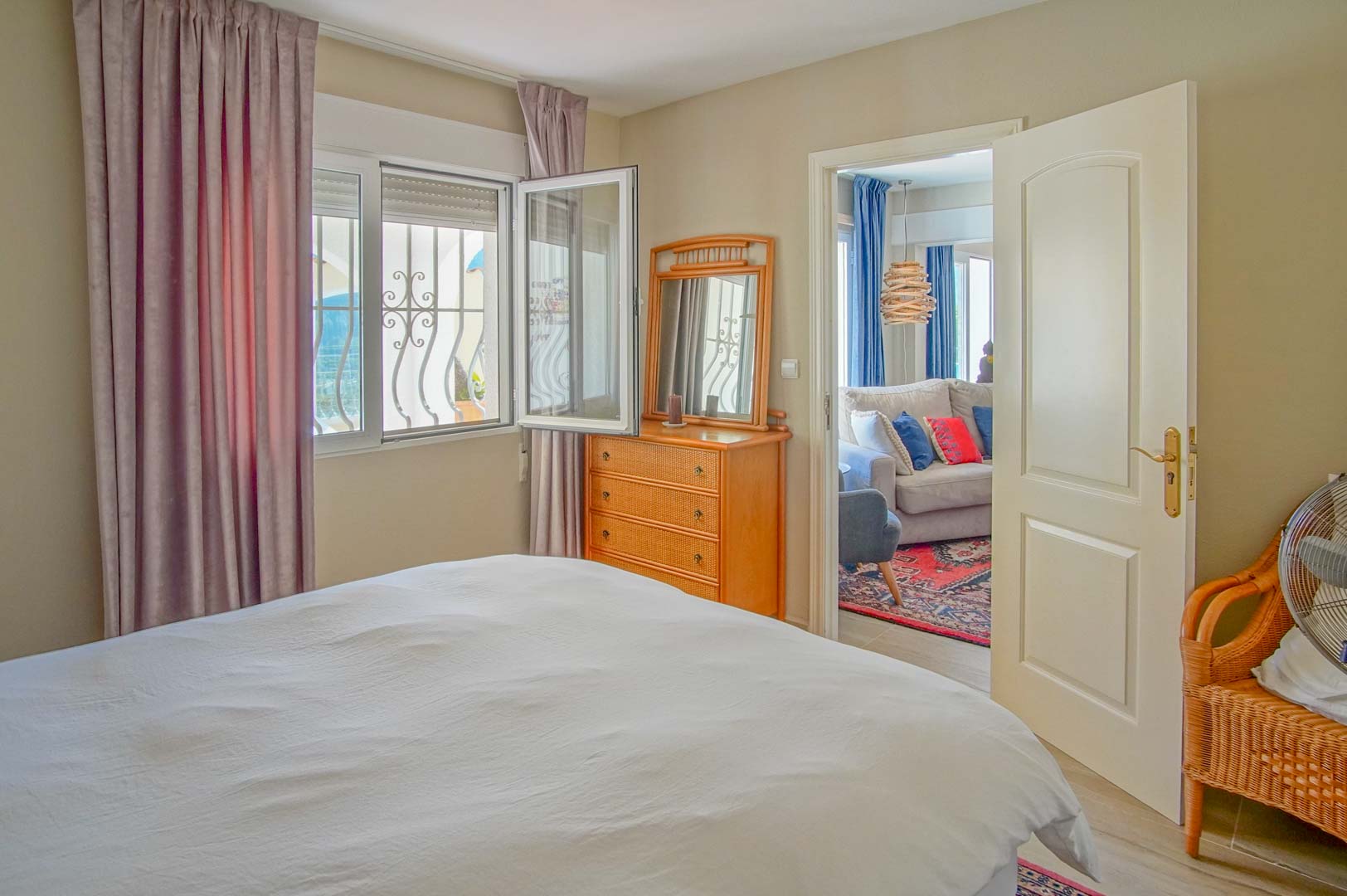 Chambre lumineuse avec commode en bois et fenêtre à barreaux décoratifs. Une porte ouverte donne sur un salon adjacent équipé d'un canapé et d'un tapis coloré.