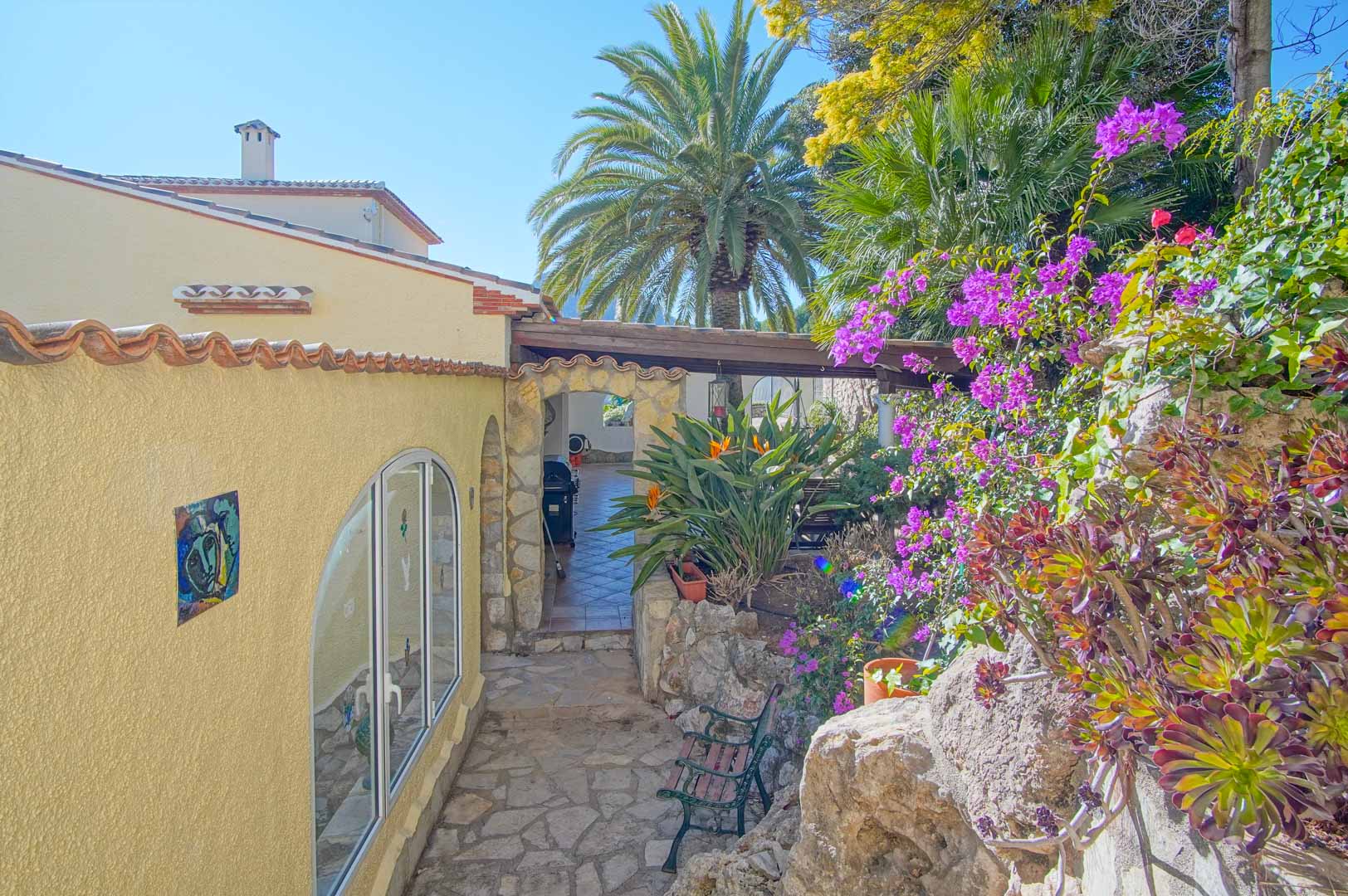 Chemin extérieur en pierre naturelle bordé de bougainvilliers, menant à une terrasse couverte via une arche en pierre authentique au cœur d'un jardin méditerranéen.