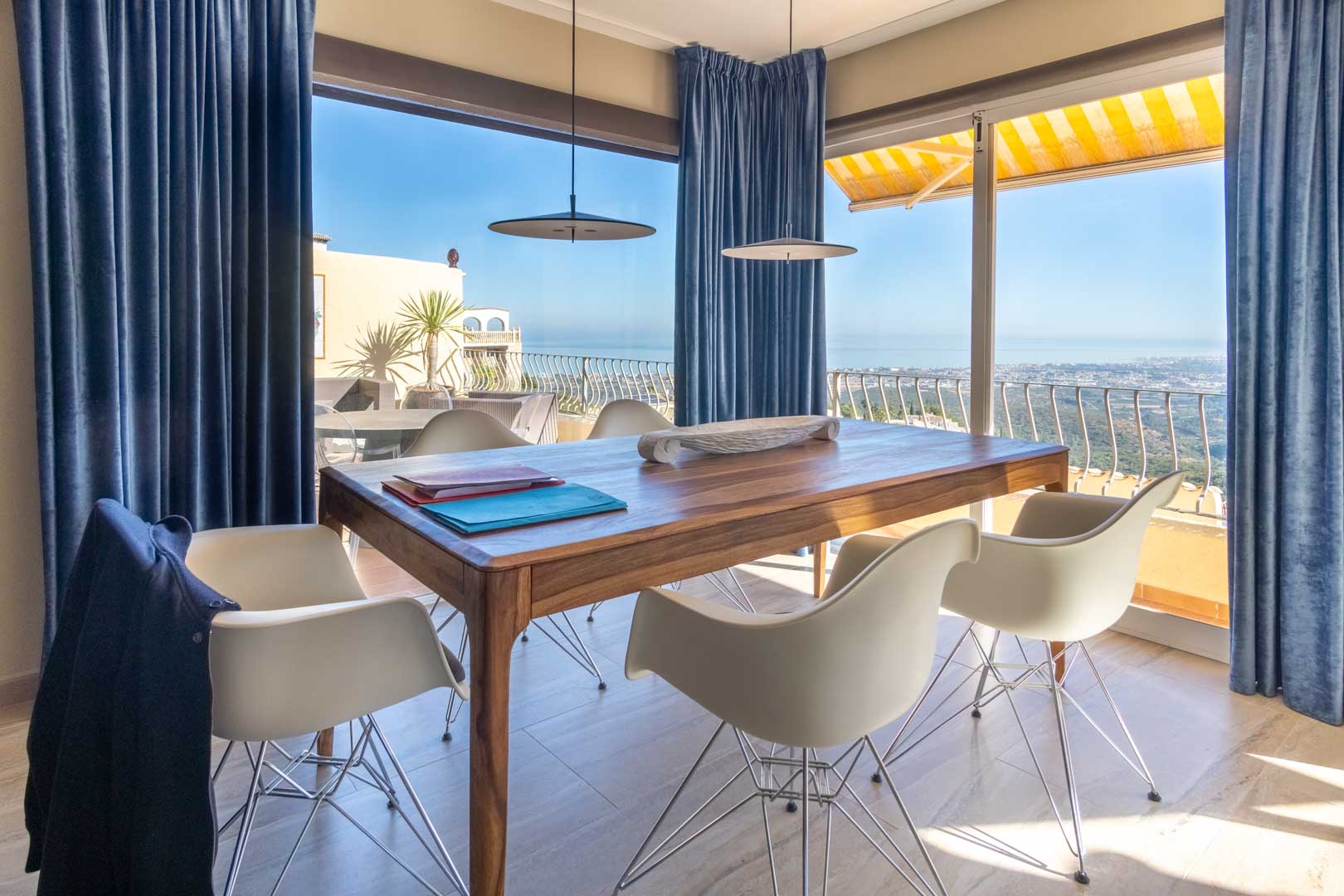 Coin repas avec table en bois massif et chaises design, doté de grandes baies vitrées offrant une vue panoramique sur la mer et la côte.