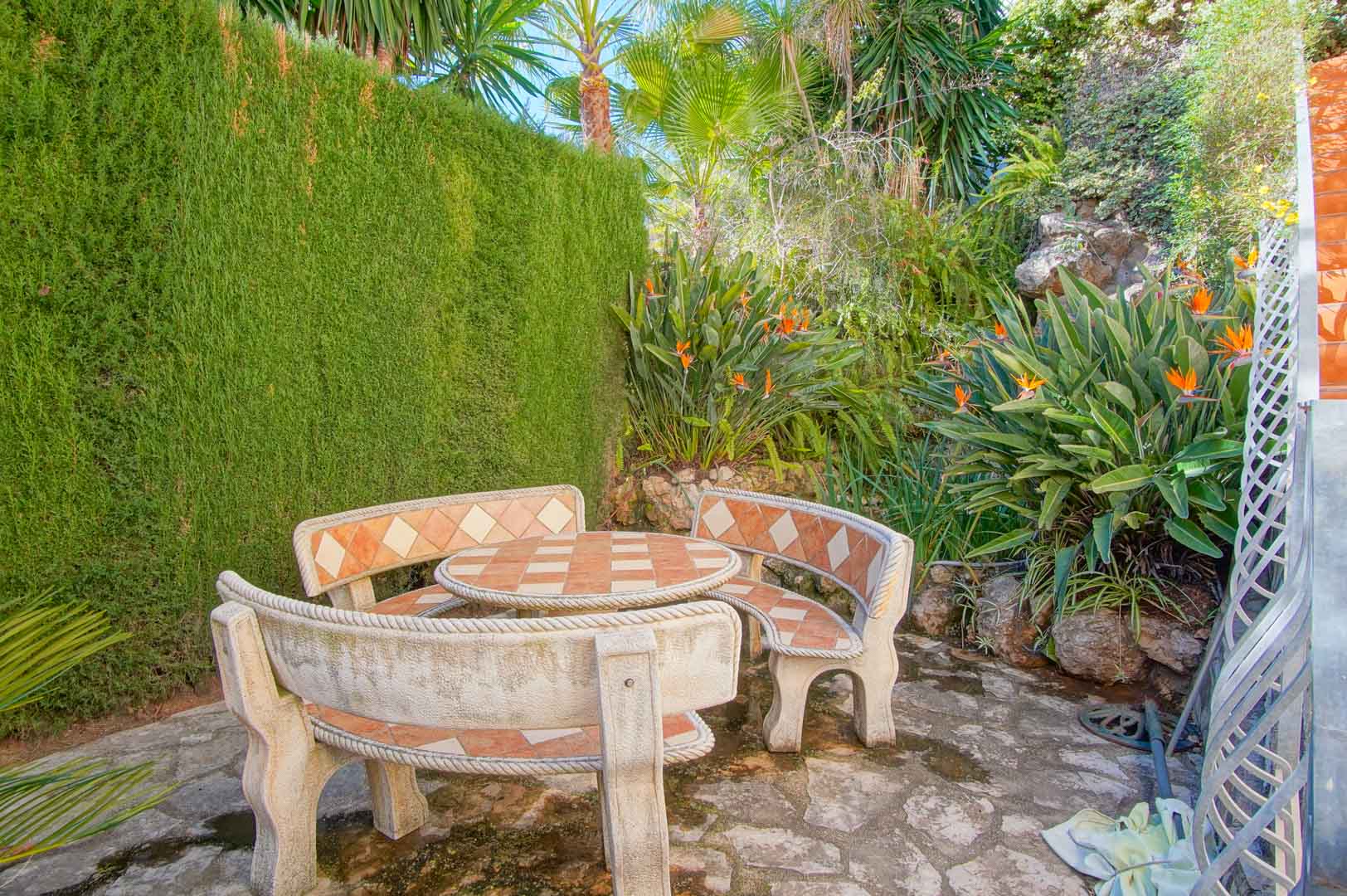 Coin salon extérieur avec table et bancs en pierre incrustés de carreaux, entouré d'une végétation luxuriante composée de palmiers et de fleurs de paradis.