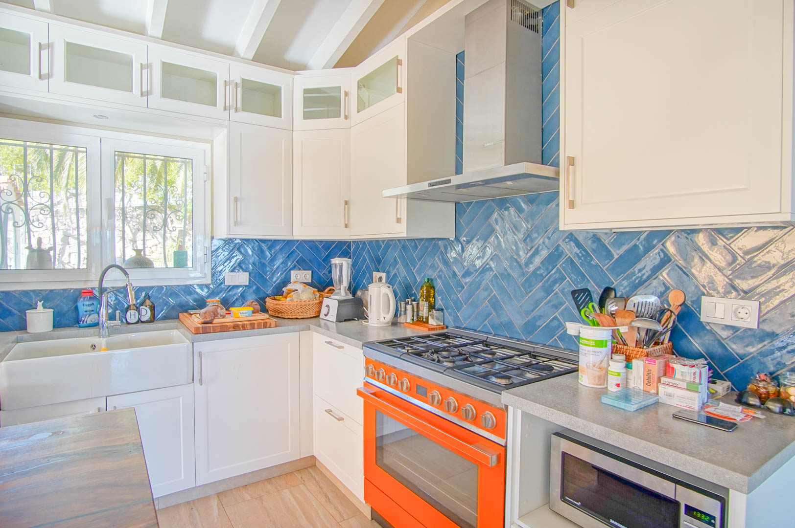 Cuisine lumineuse avec crédence en chevrons bleus, plan de travail gris et piano de cuisson orange. Évier timbre d'office et poutres apparentes blanches au plafond.