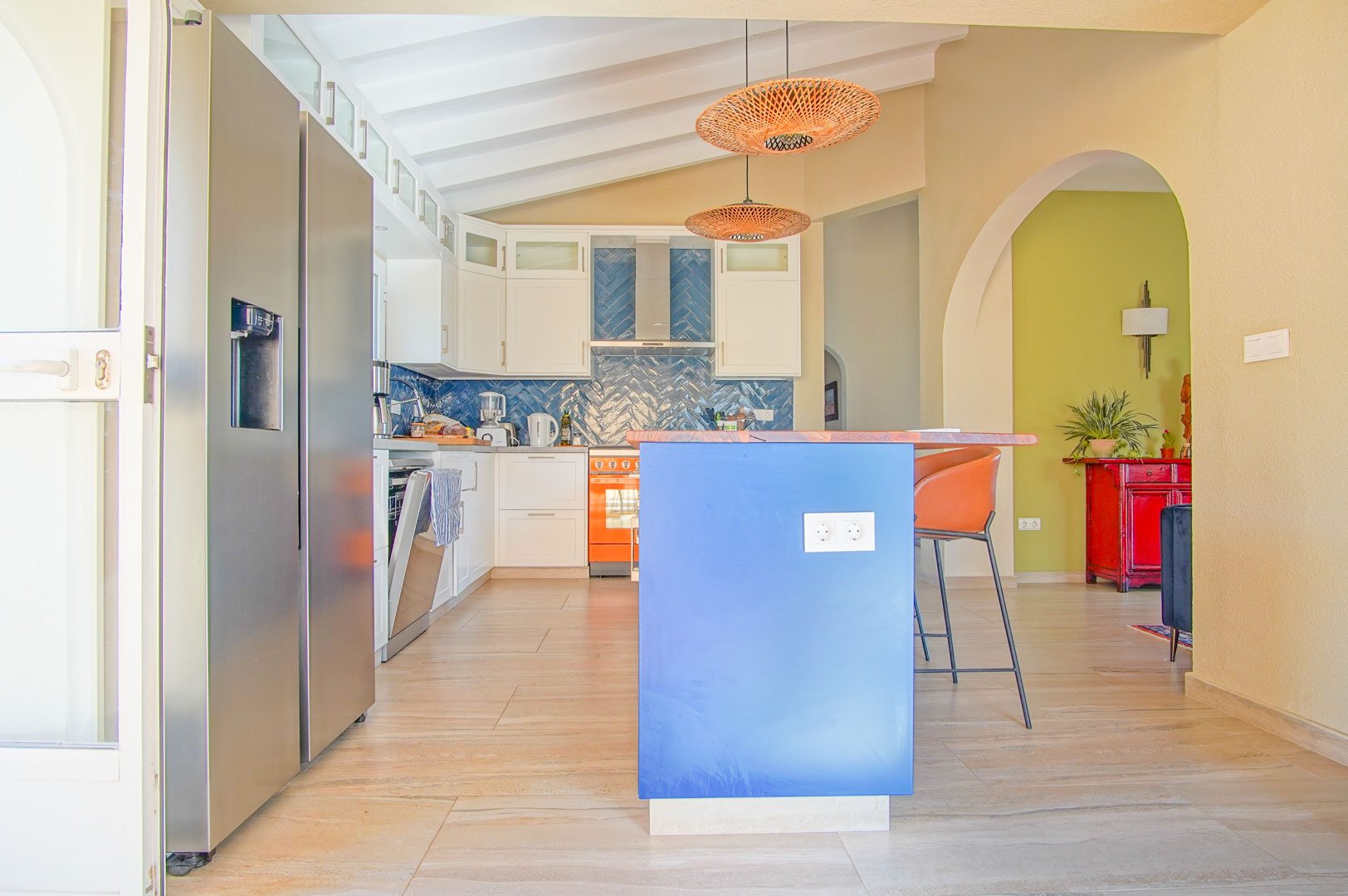 Cuisine moderne avec îlot bleu, plan de travail en bois et crédence en chevrons. Plafond incliné à poutres blanches, luminaires en rotin et électroménagers en inox.