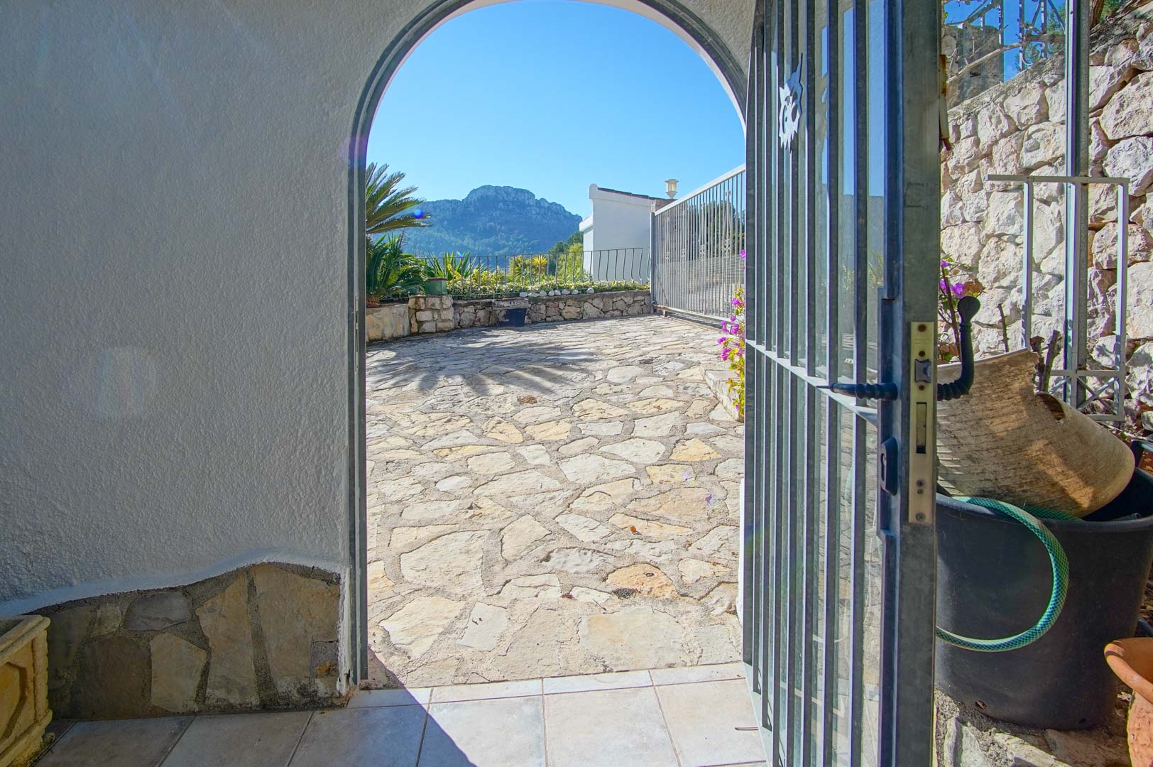 Entrée en arche menant à une terrasse en pierre naturelle avec vue sur la montagne, entourée de murs en pierre et d'une clôture métallique.