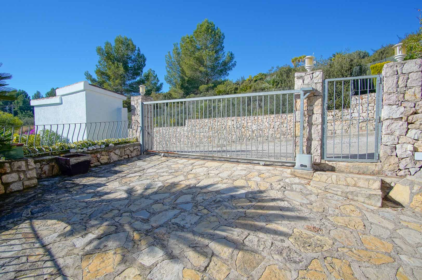 Entrée principale avec portail coulissant automatique et portillon, entourée de murs en pierre naturelle et d'un sol en dalles irrégulières.