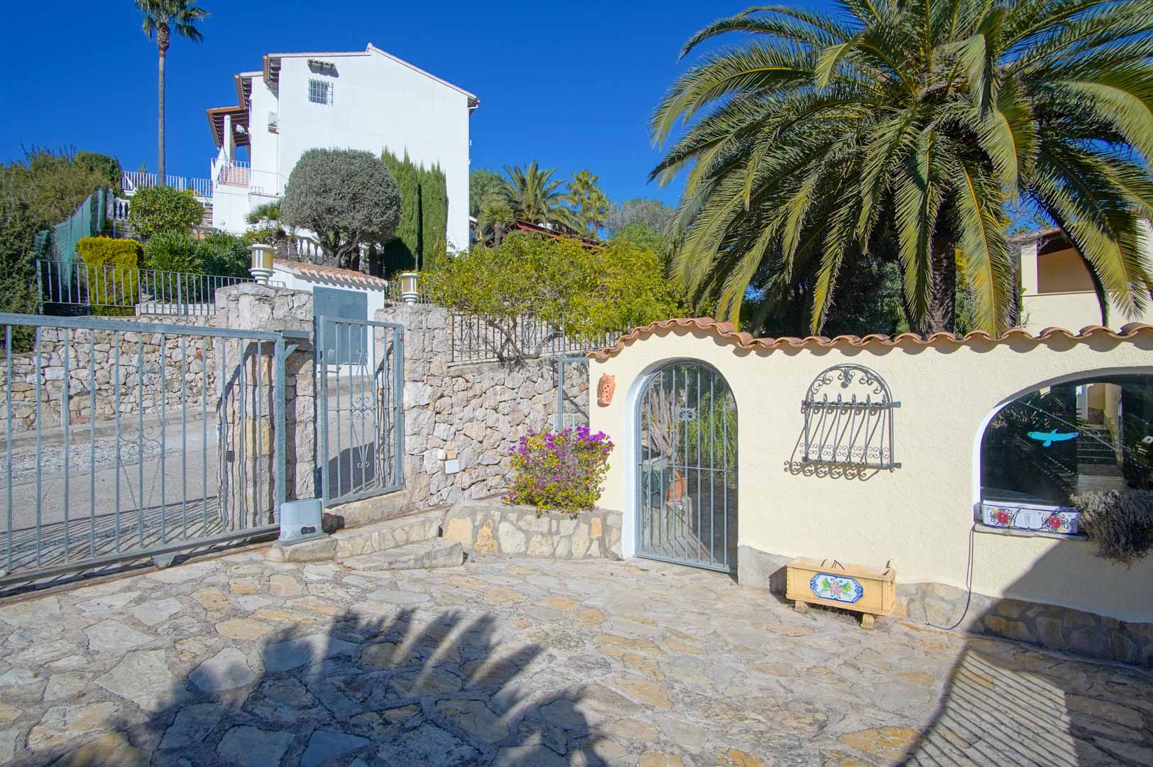Entrée principale avec portail coulissant en métal, murs en pierre naturelle et porte piétonne voûtée, entourée de palmiers et d'une cour pavée.