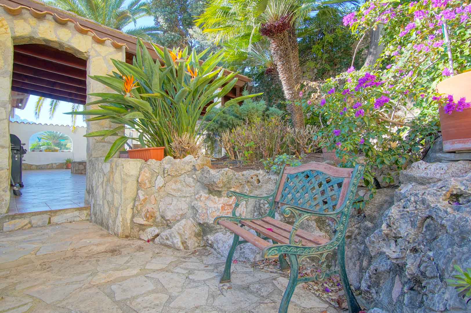 Espace de repos extérieur avec banc en fer forgé, murs en pierre et plantes méditerranéennes, situé à côté d'une terrasse couverte aux poutres apparentes.
